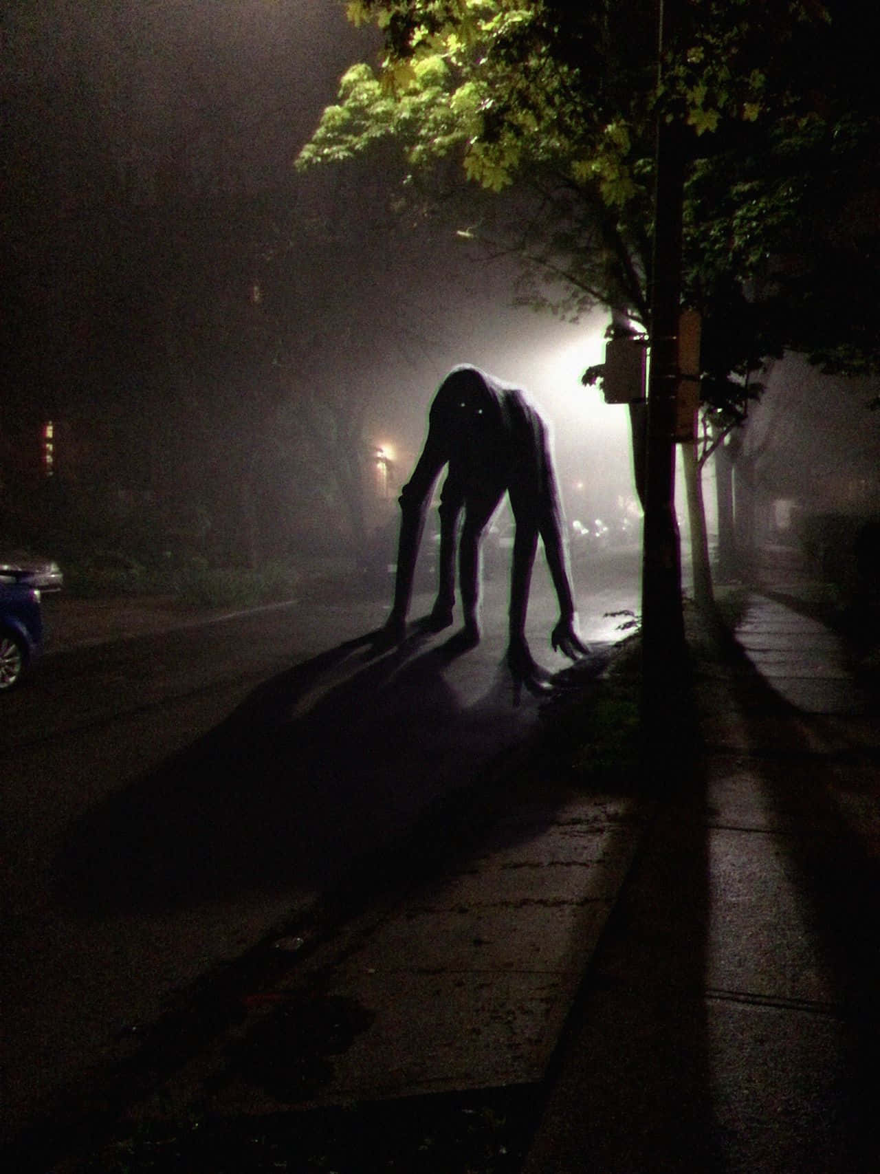 詭異的GIF 桌布 壁紙 — 一個高聳的陰影生物在夜間蹲在霧濛濛的城市人行道上，被街燈照亮，投下長長的詭異剪影。