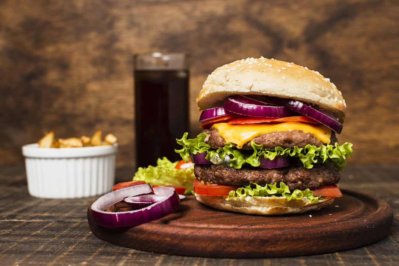 fast food 桌布 壁紙 — 一個高聳的雙層起司漢堡，配上新鮮生菜、番茄和洋蔥，放在木板上，旁邊有薯條和深色汽水。