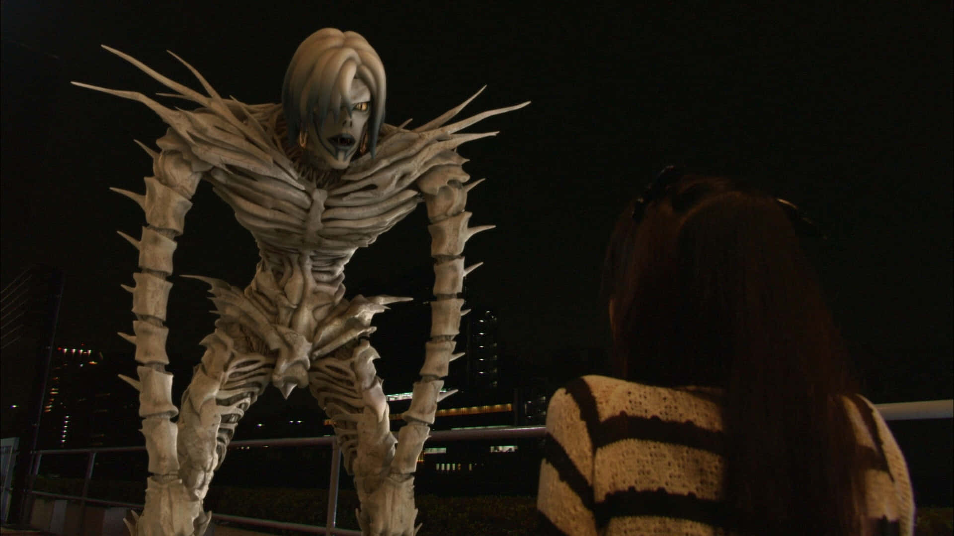 Death Note Rem 桌布 壁紙 — 一個高聳的骷髏動畫怪物，帶有尖刺突起，在夜晚的城市屋頂上與一個女孩對峙。