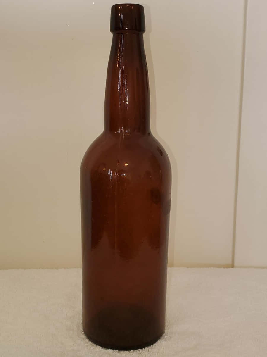 Old Bottles 桌布 壁紙 — 一個高身琥珀色玻璃瓶，長頸，直立於白色紋理表面上，背景為素色牆壁。