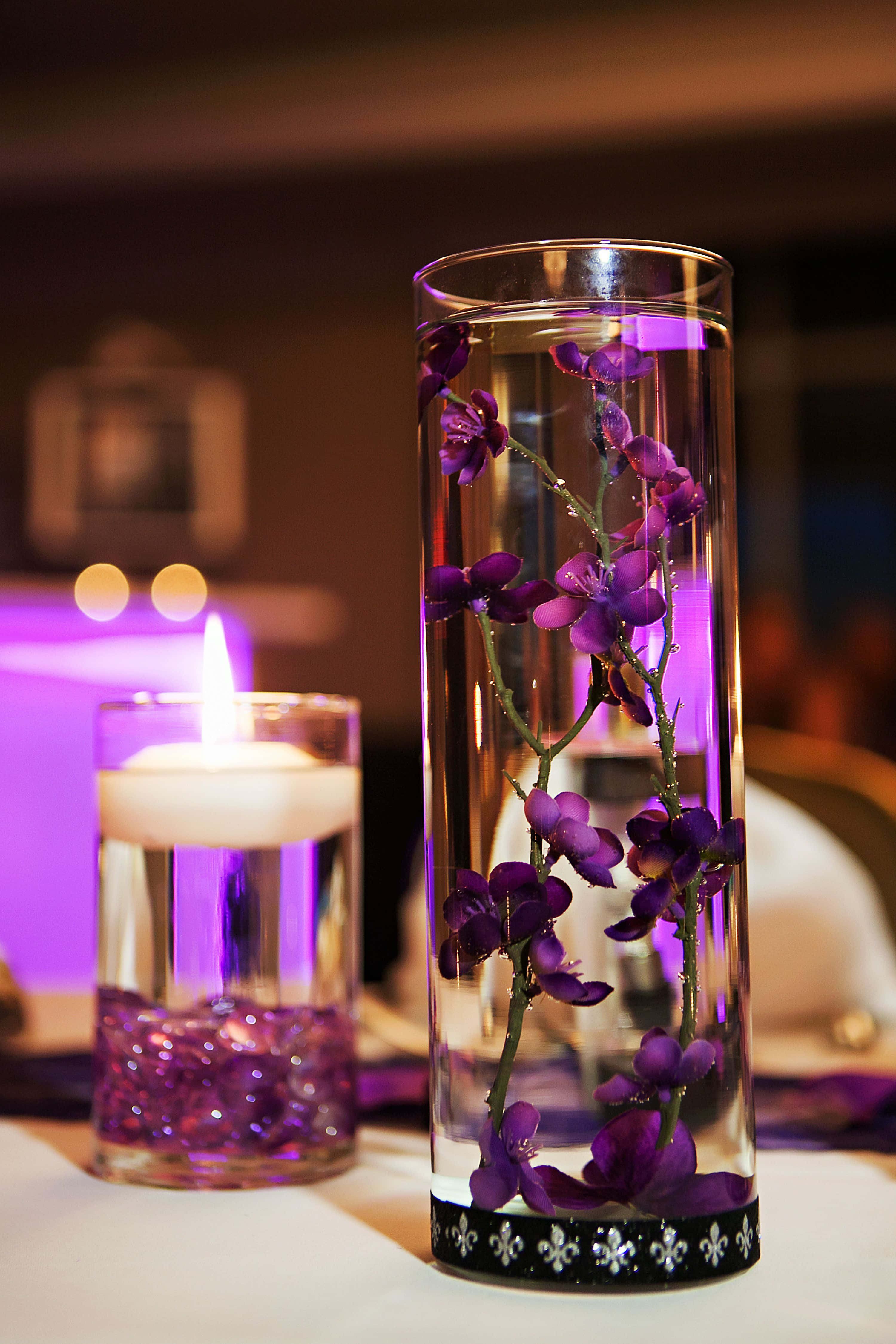 Centerpiece 桌布 壁紙 — 一個高高的玻璃花瓶，裝滿水和紫色蘭花，旁邊放著一個點燃的浮動蠟燭，帶有紫色珠子，在柔和光線的房間裡。