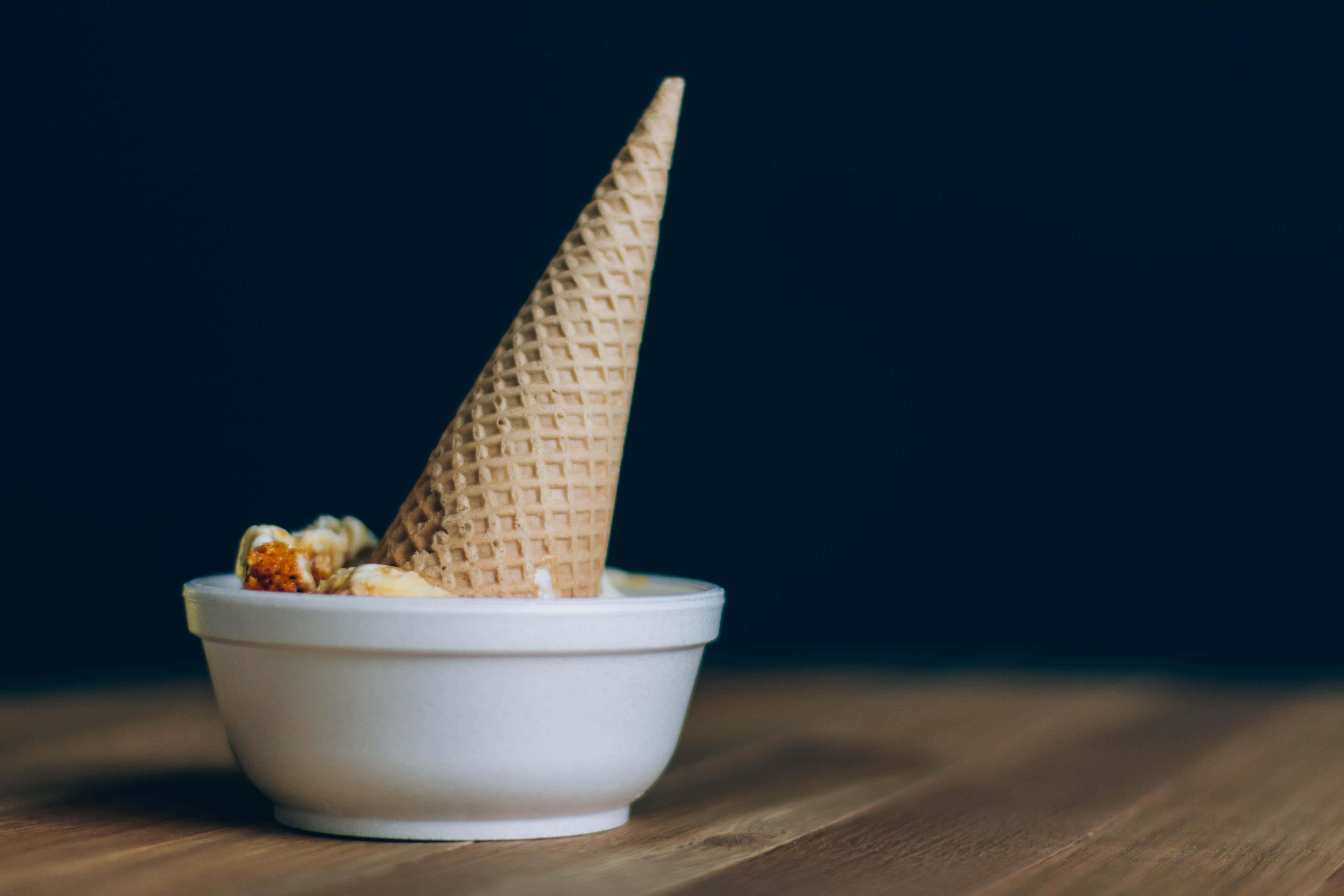 Ice Cream 桌布 壁紙 — 一個鬆餅筒直立於白色碗中，木質表面上散落著冰淇淋碎塊，背景為深色。