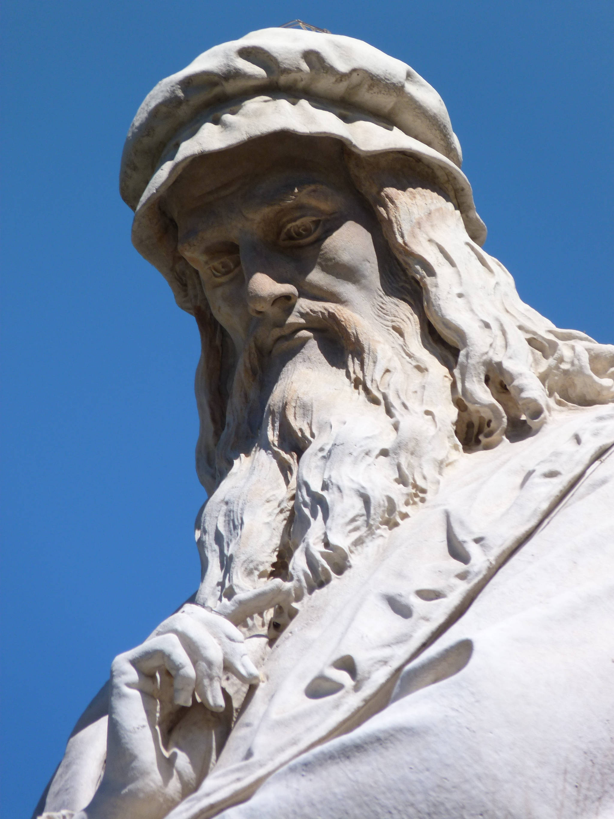 Leonardo Da Vinci 桌布 壁紙 — 一個鬍鬚男人戴帽子的風化白色大理石雕像特寫，背景是清澈的藍天。