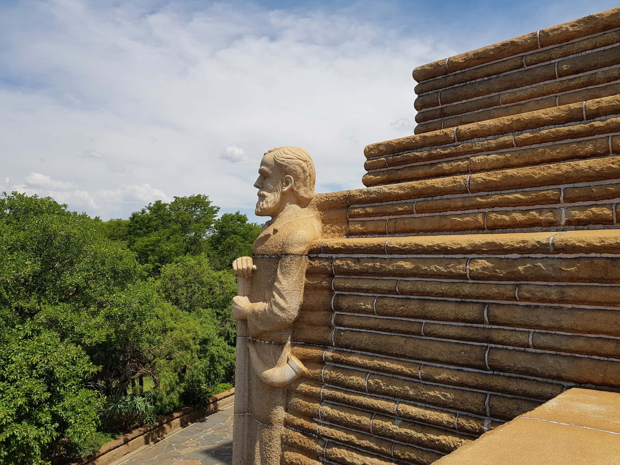 statue 桌布 壁紙 — 一個鬍鬚男人的石像站在堆疊的木牆旁邊，背景是部分多雲的天空和茂密的綠樹。