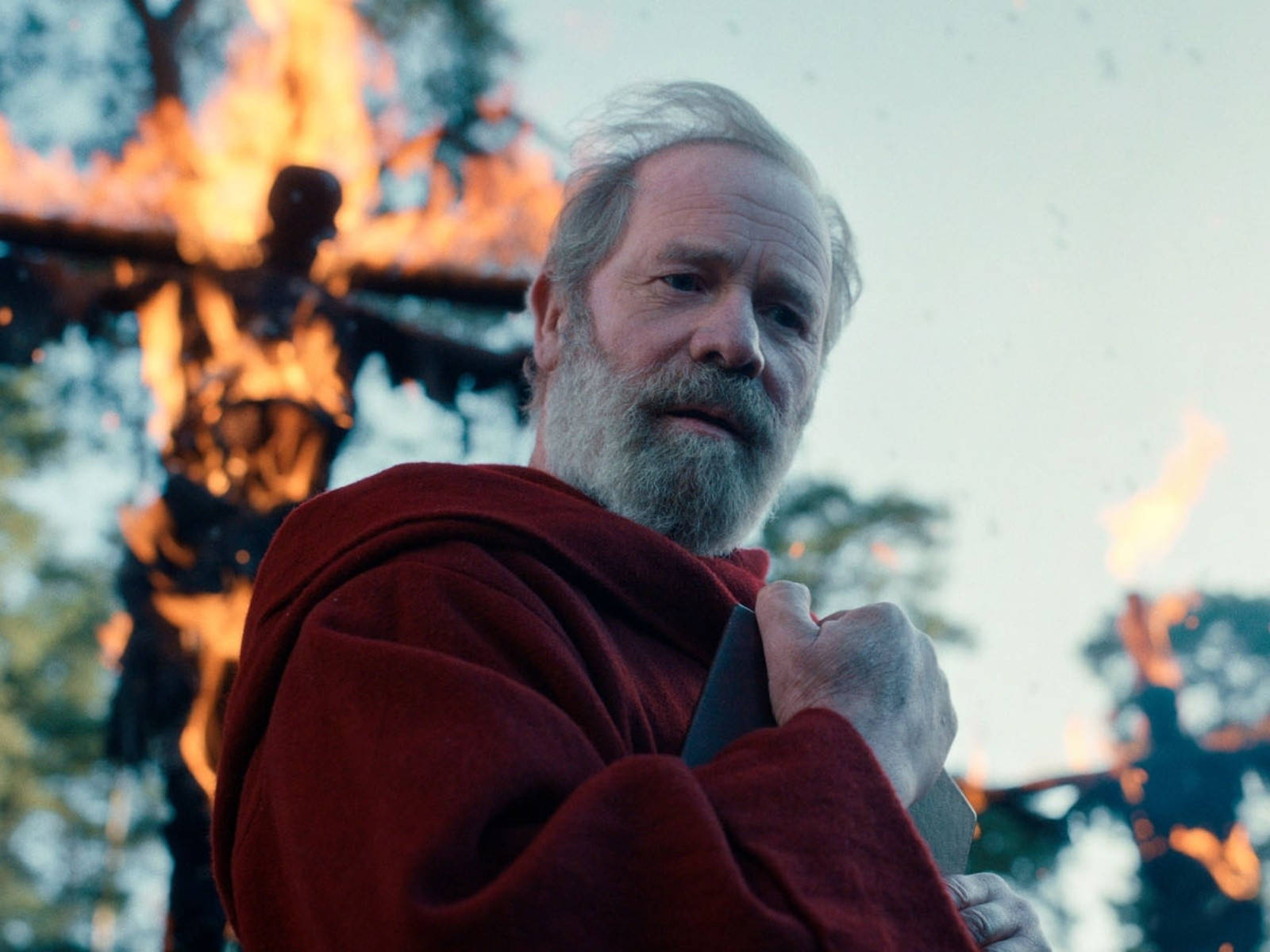 Netflix 桌布 壁紙 — 一個鬍鬚男人身穿紅色長袍，在黃昏的森林中站在一個燃燒的木製肖像前，手持一把刀。