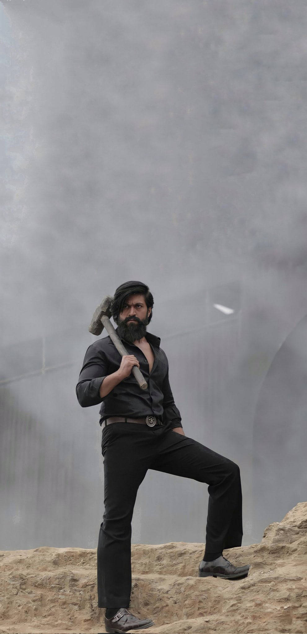 Bollywood Hero 桌布 壁紙 — 一個鬍鬚男人身穿黑色衣服，自信地站在岩石地形上，手持錘子，背景是霧氣瀰漫的工業場景。