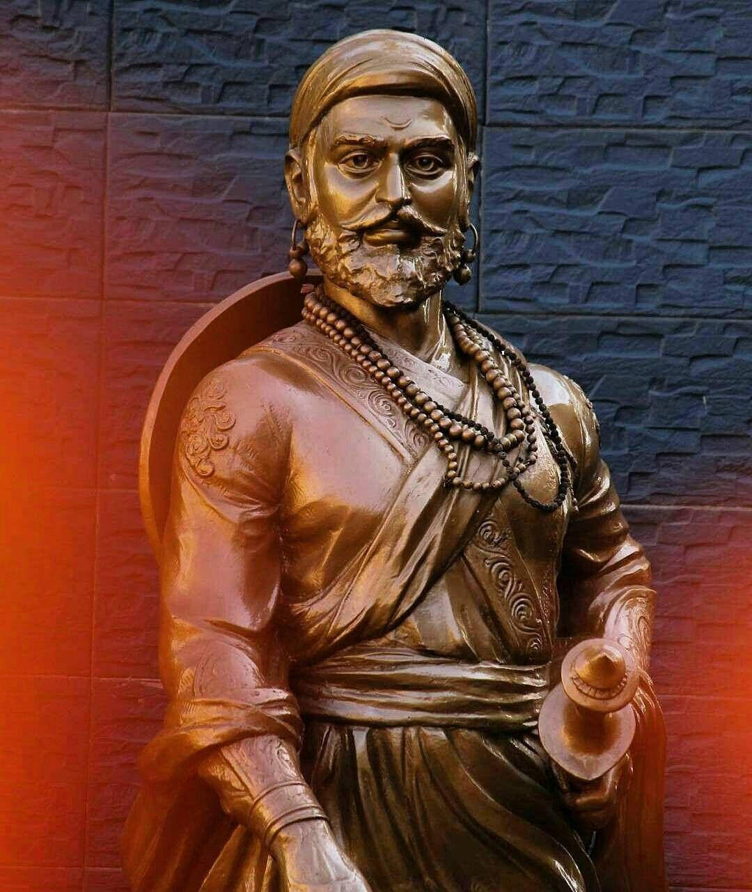 Chhatrapati Shivaji Maharaj 桌布 壁紙 — 一個鬍鬚男子穿著傳統服飾，手持杯子，面向有紋理的牆壁，牆上有溫暖的紅色和冷藍色燈光。