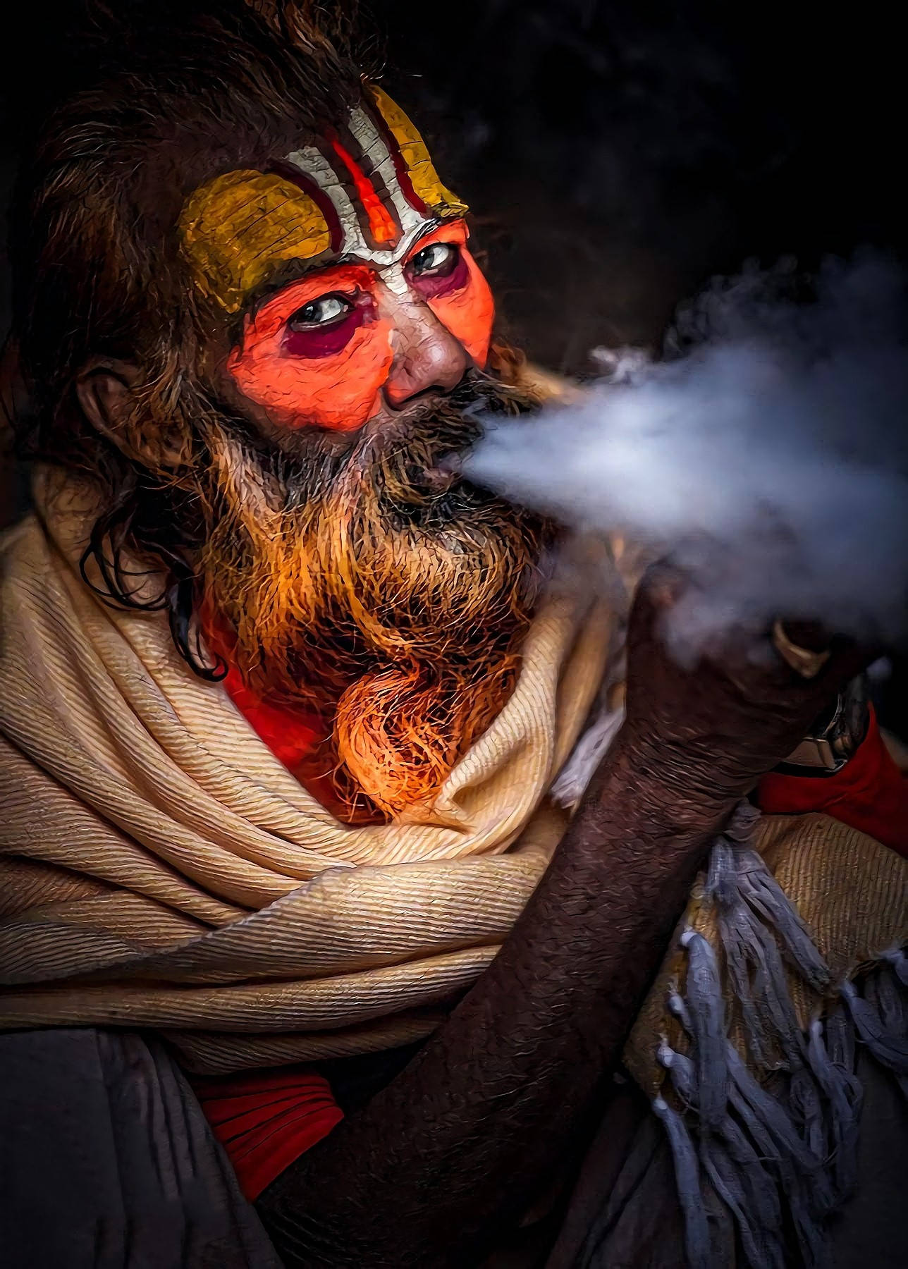 Aghori 桌布 壁紙 — 一個鬍鬚男的特寫，他臉上塗有鮮豔的橙色和金色顏料，在黑暗背景前呼出煙霧。
