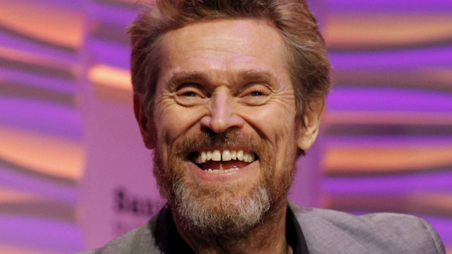 Willem Dafoe 桌布 壁紙 — 一個鬍鬚男的特寫，在充滿活力的紫色和橙色條紋背景上露出燦爛的笑容。
