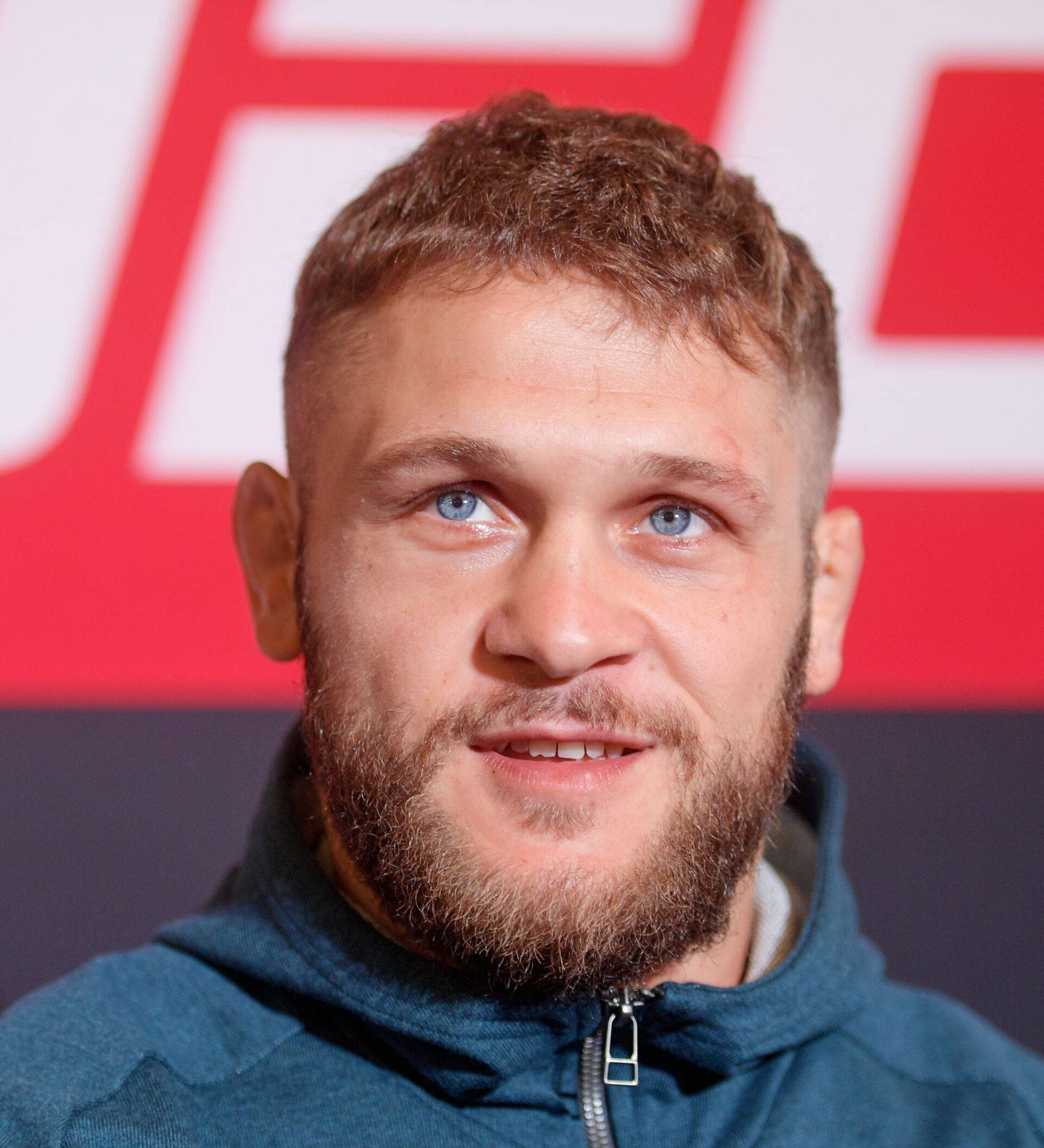 UFC 桌布 壁紙 — 一個鬍鬚男的近距離肖像，擁有引人注目的藍色眼睛，在紅白相間的UFC背景下微微微笑。
