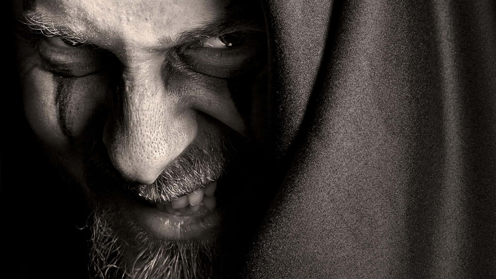 Face 桌布 壁紙 — 一個鬍鬚男的面部特寫，半邊臉被布料遮蓋，在戲劇性的明暗對比光線下，瞇著眼睛露出邪惡的笑容。