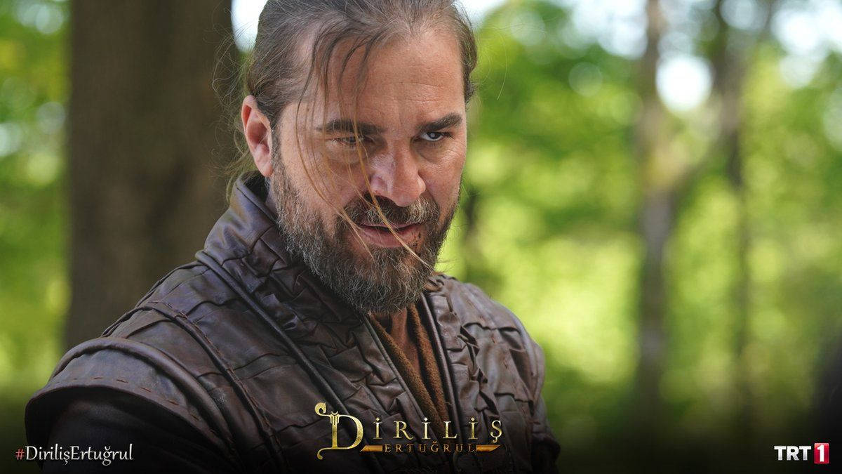 Ertugrul 桌布 壁紙 — 一個鬍鬚男穿著皮甲，透過陽光照耀的森林樹葉凝視著。