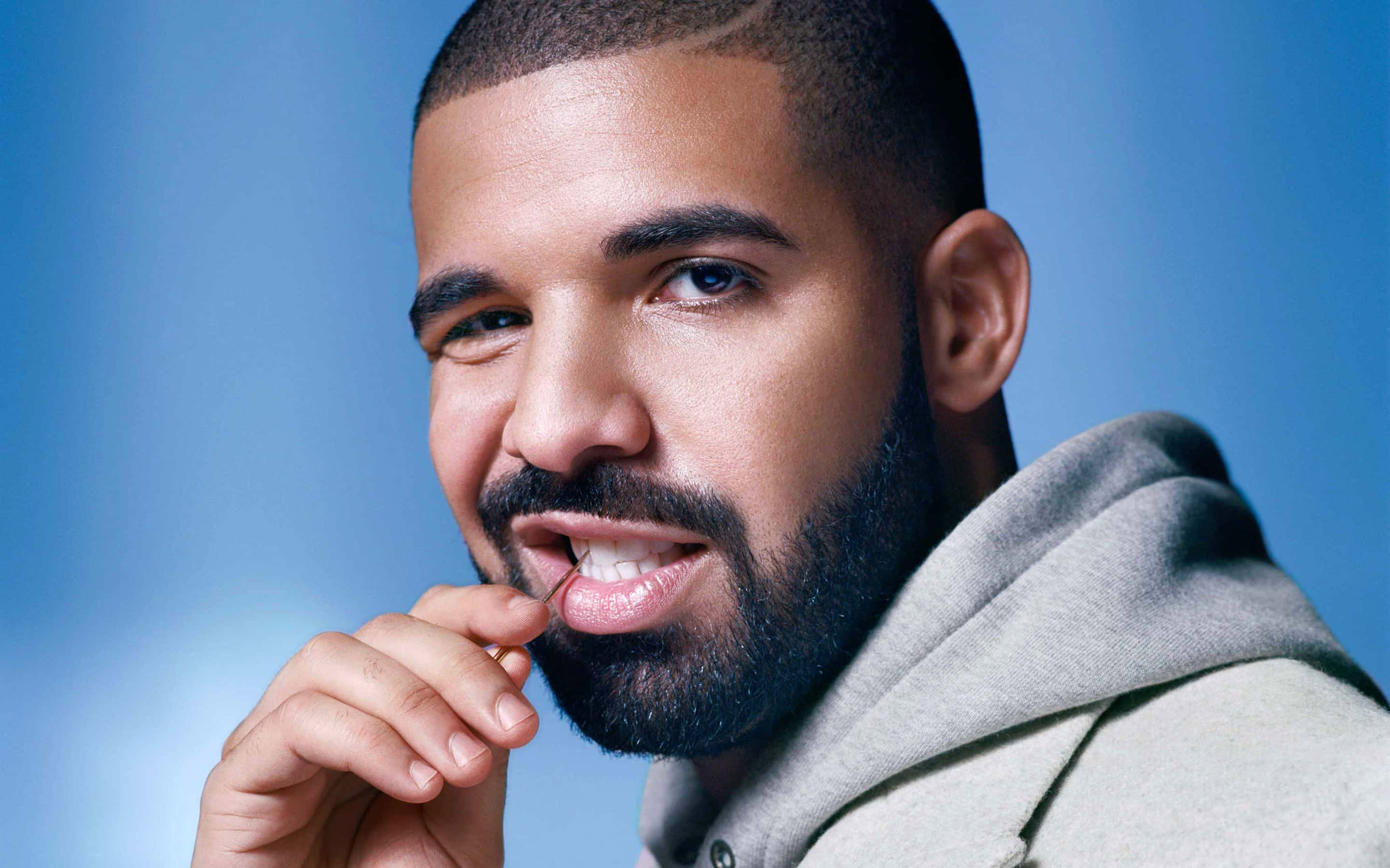 Aesthetic Drake 桌布 壁紙 — 一個鬍鬚男穿著連帽衫，在柔和藍色背景前微笑觸摸嘴唇的特寫肖像。