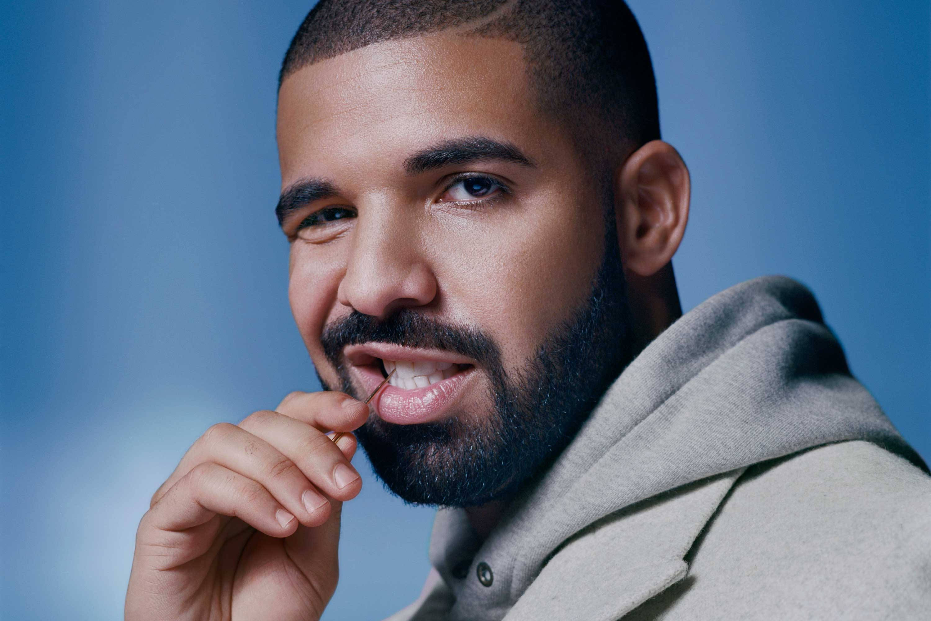 Drake 桌布 壁紙 — 一個鬍鬚短髮男子的特寫肖像，微笑著將一個小物件靠近嘴邊，背景是柔和的藍色。