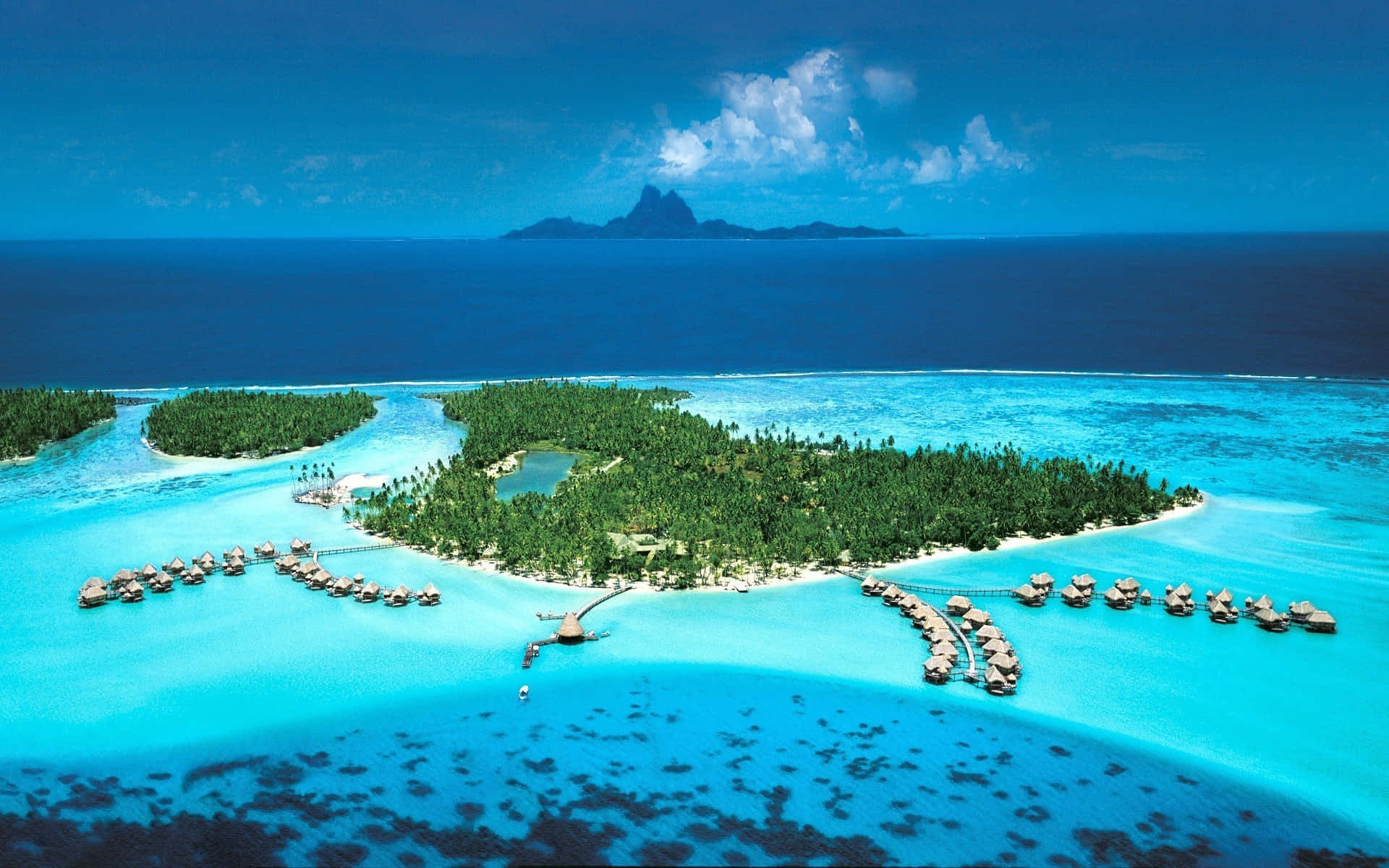 Bora Bora 桌布 壁紙 — 一個鬱鬱蔥蔥的熱帶島嶼的鳥瞰圖，島上有水上平房，坐落在碧綠色的潟湖中，周圍是深藍色的海洋和遠處的火山峰。