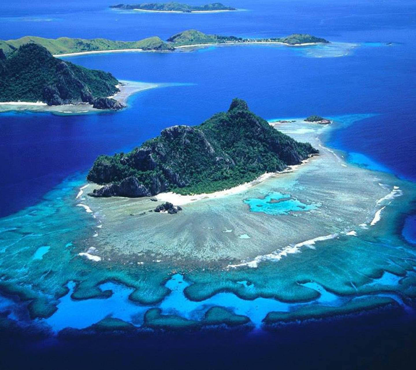 Fiji 桌布 壁紙 — 一個鬱鬱蔥蔥的綠色島嶼的鳥瞰圖，被充滿活力的綠松石珊瑚礁和深藍色海水包圍。