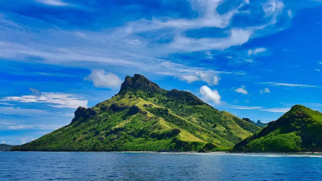 自然 桌布 壁紙 — 一個鬱鬱蔥蔥的綠色火山島從平靜的藍色海水中陡峭升起，上方是充滿活力的藍天和縷縷白雲。