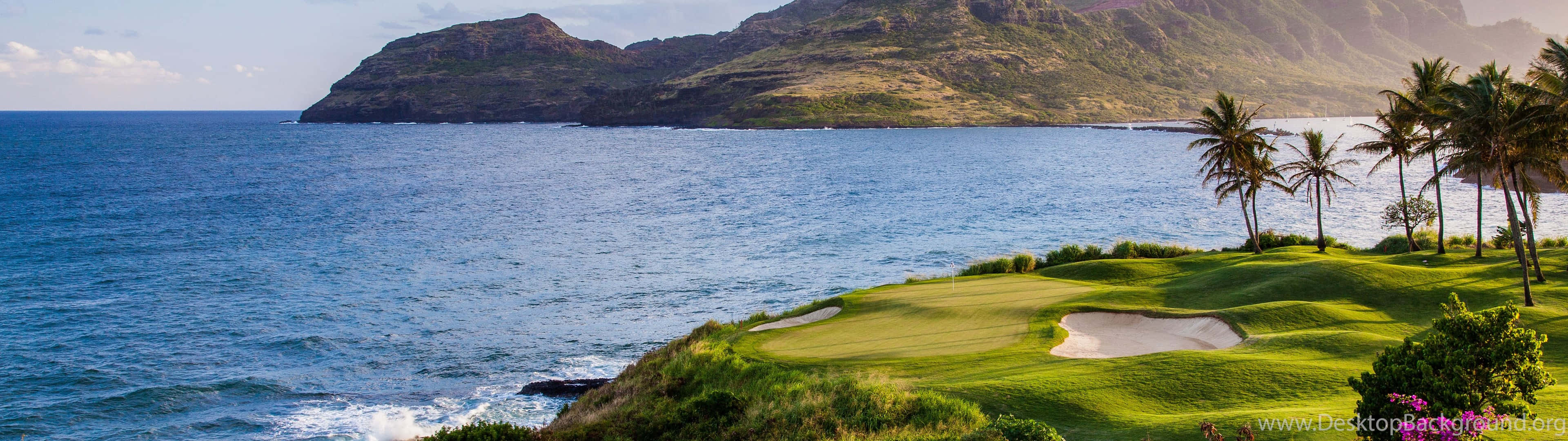 Golf 3840x1080 桌布 壁紙 — 一個鬱鬱蔥蔥的高爾夫球場緊貼懸崖，俯瞰著碧綠色的海洋，遠處有棕櫚樹和崎嶇的山脈。