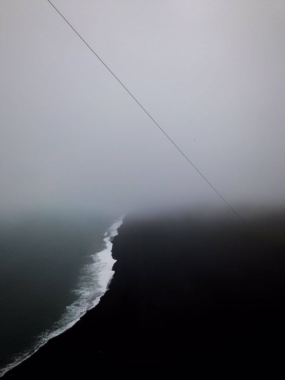 一個霧氣瀰漫的海岸場景，黑暗的海灘和海洋波浪在灰色天空下，帶有對角線纜繩。 — 人像動態 壁紙