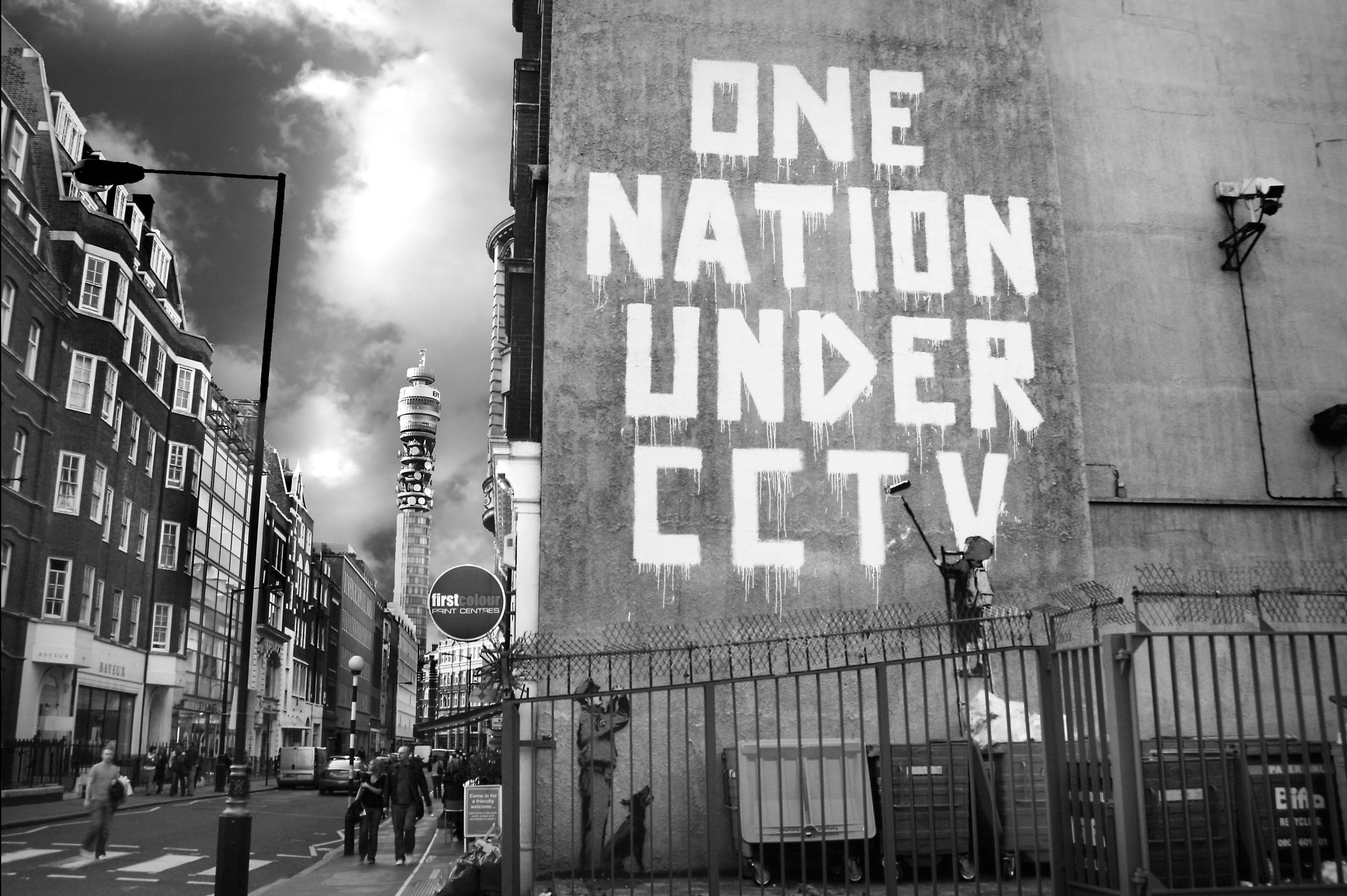 Banksy 桌布 壁紙 — 一個鮮明的黑白城市場景，在混凝土牆上有一幅塗鴉壁畫，上面寫著「ONE NATION UNDER CCTV」，旁邊是帶有監控攝像頭的圍欄。