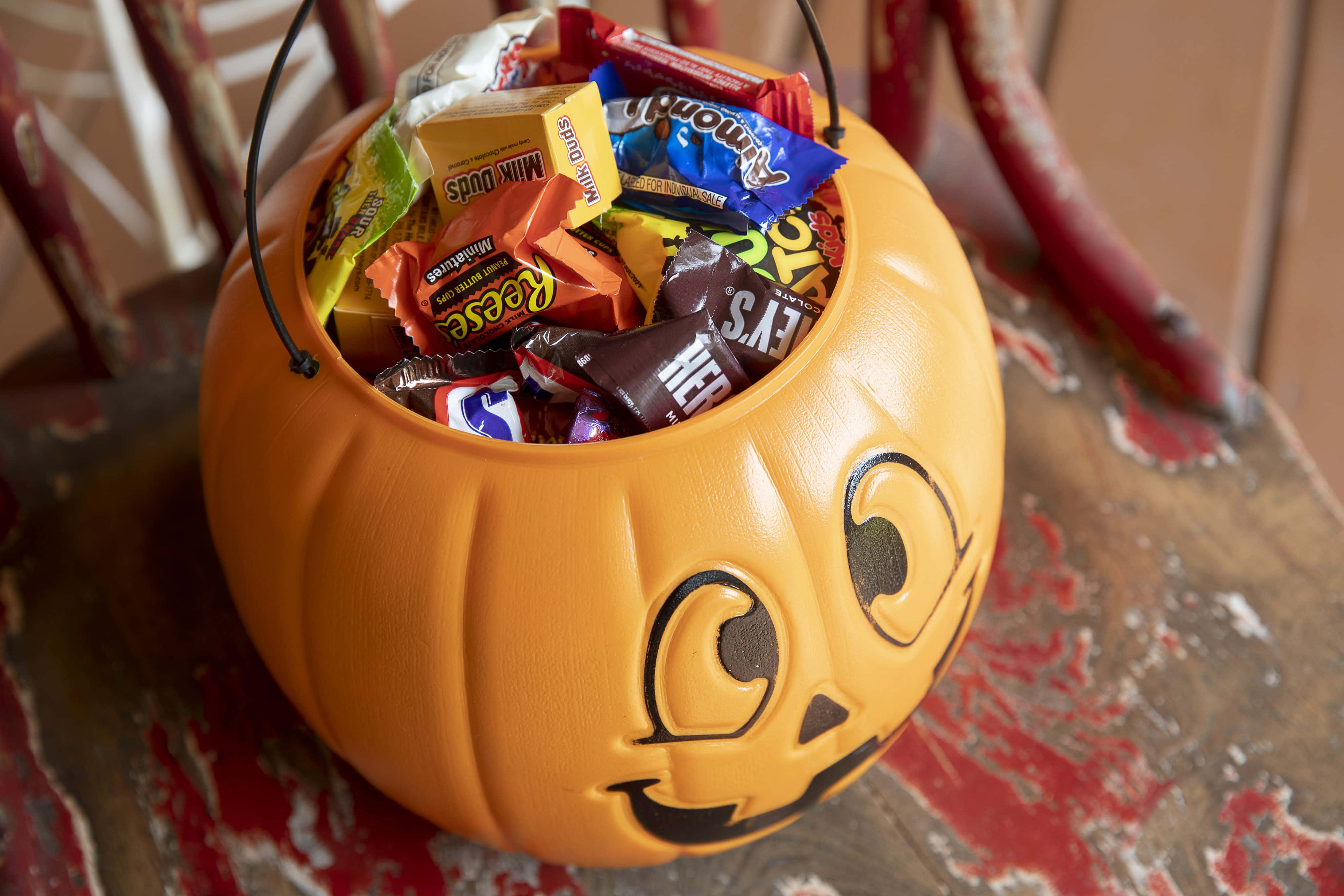 Halloween Treats 桌布 壁紙 — 一個鮮橙色南瓜形狀的萬聖節糖果桶，裝滿了各種色彩繽紛的糖果，放在一張風化的紅色木椅上。