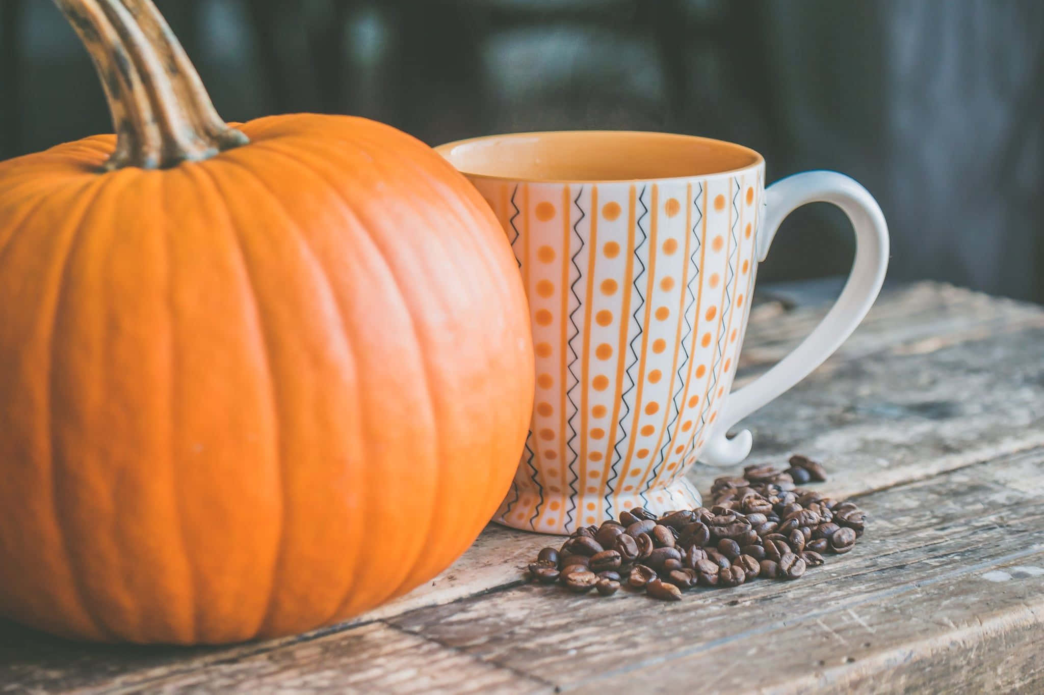 Pumpkin 桌布 壁紙 — 一個鮮橙色的南瓜放在一個有圖案的杯子和散落的咖啡豆旁邊，置於一張鄉村風格的木桌上。