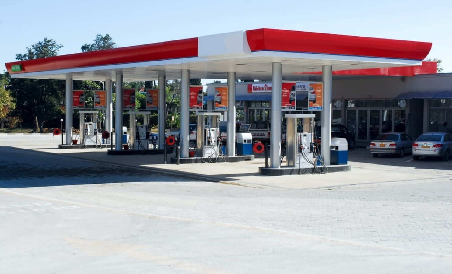 Petrol Station 桌布 壁紙 — 一個鮮紅色和白色的加油站頂篷在晴朗的藍天下遮蓋著多個油泵，附近停有汽車。