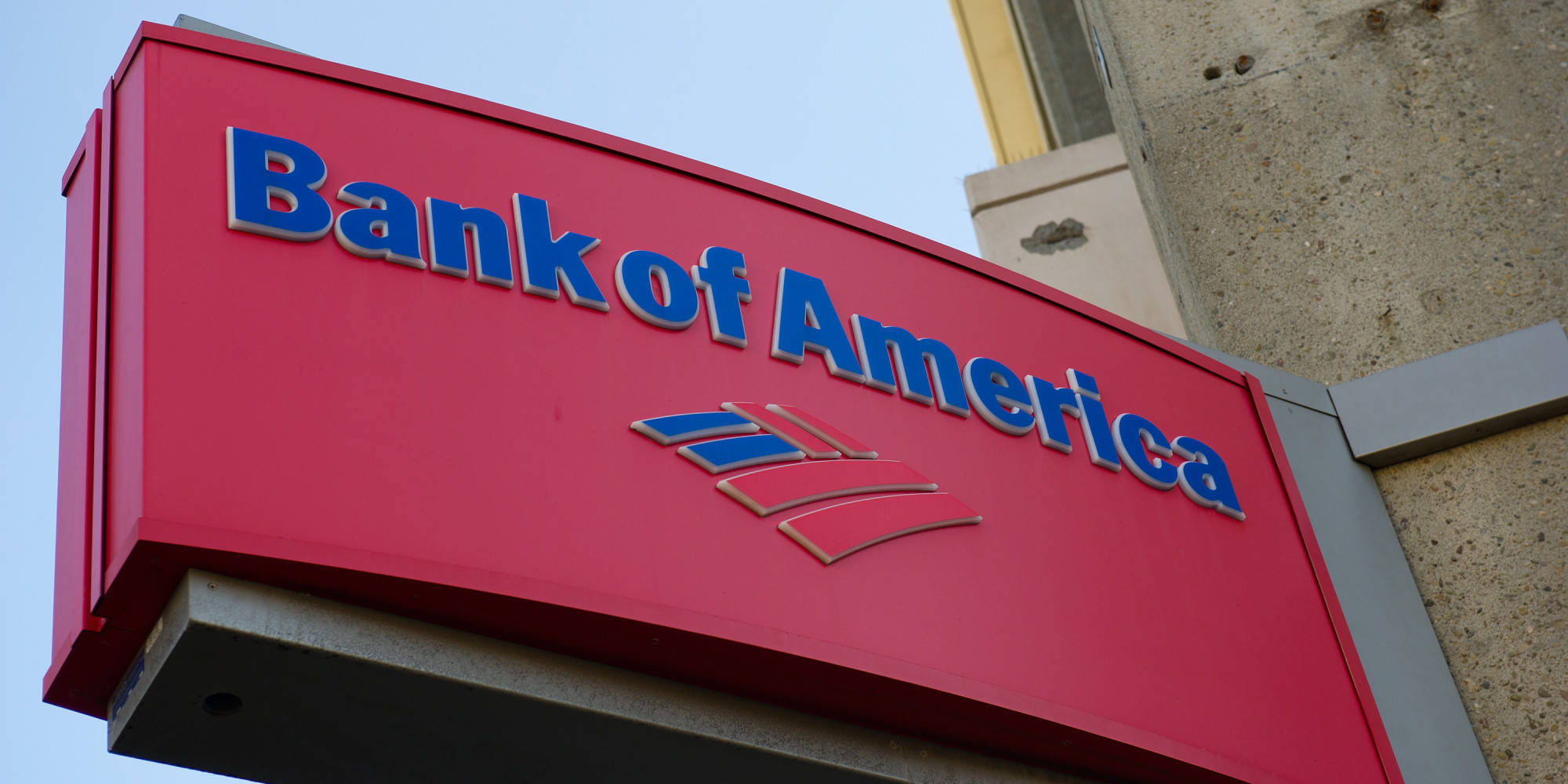Bank 桌布 壁紙 — 一個鮮紅色的Bank of America招牌，帶有藍色字體和標誌，安裝在晴朗天空下的混凝土建築物上。