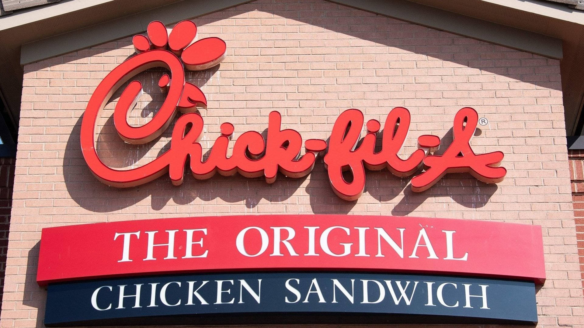 chick 桌布 壁紙 — 一個鮮紅色的Chick-fil-A標誌，帶有粗體字，安裝在磚砌建築上，在晴朗的日光下。