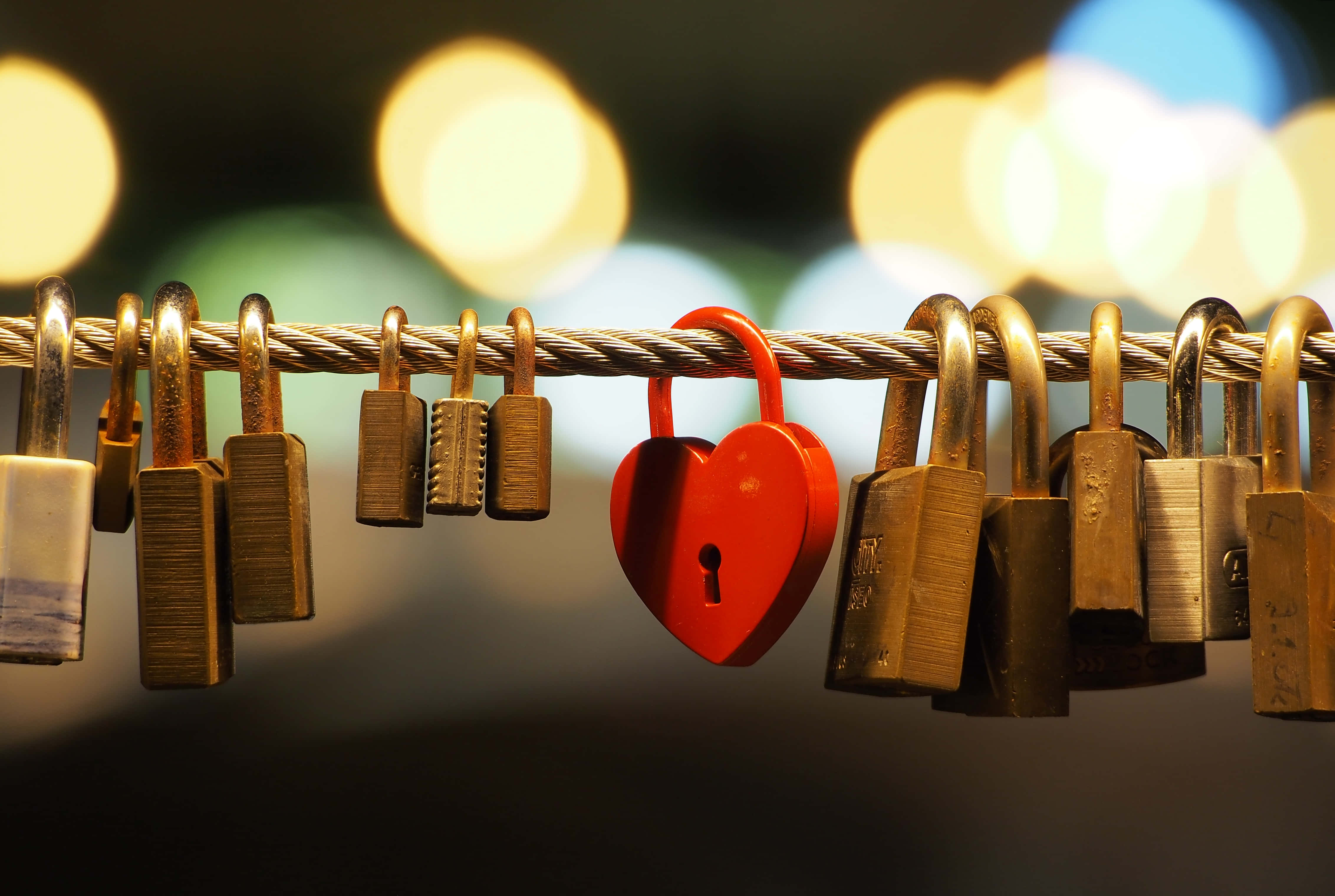 Locks 桌布 壁紙 — 一個鮮紅色的心形掛鎖在掛在電纜上的風化黃銅鎖中脫穎而出，背景是散景的城市夜景。