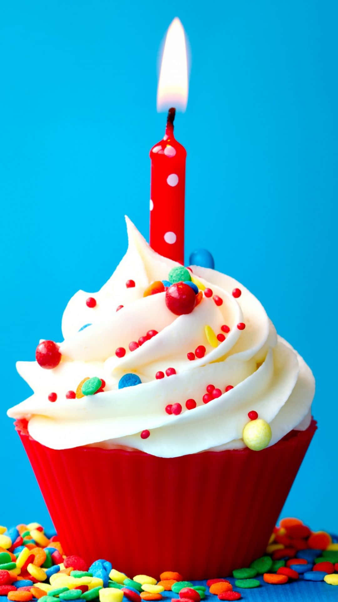 Birthday 桌布 壁紙 — 一個鮮紅色的杯子蛋糕，上面有白色糖霜和一根點燃的蠟燭，周圍是色彩繽紛的糖粒，背景是明亮的藍色。