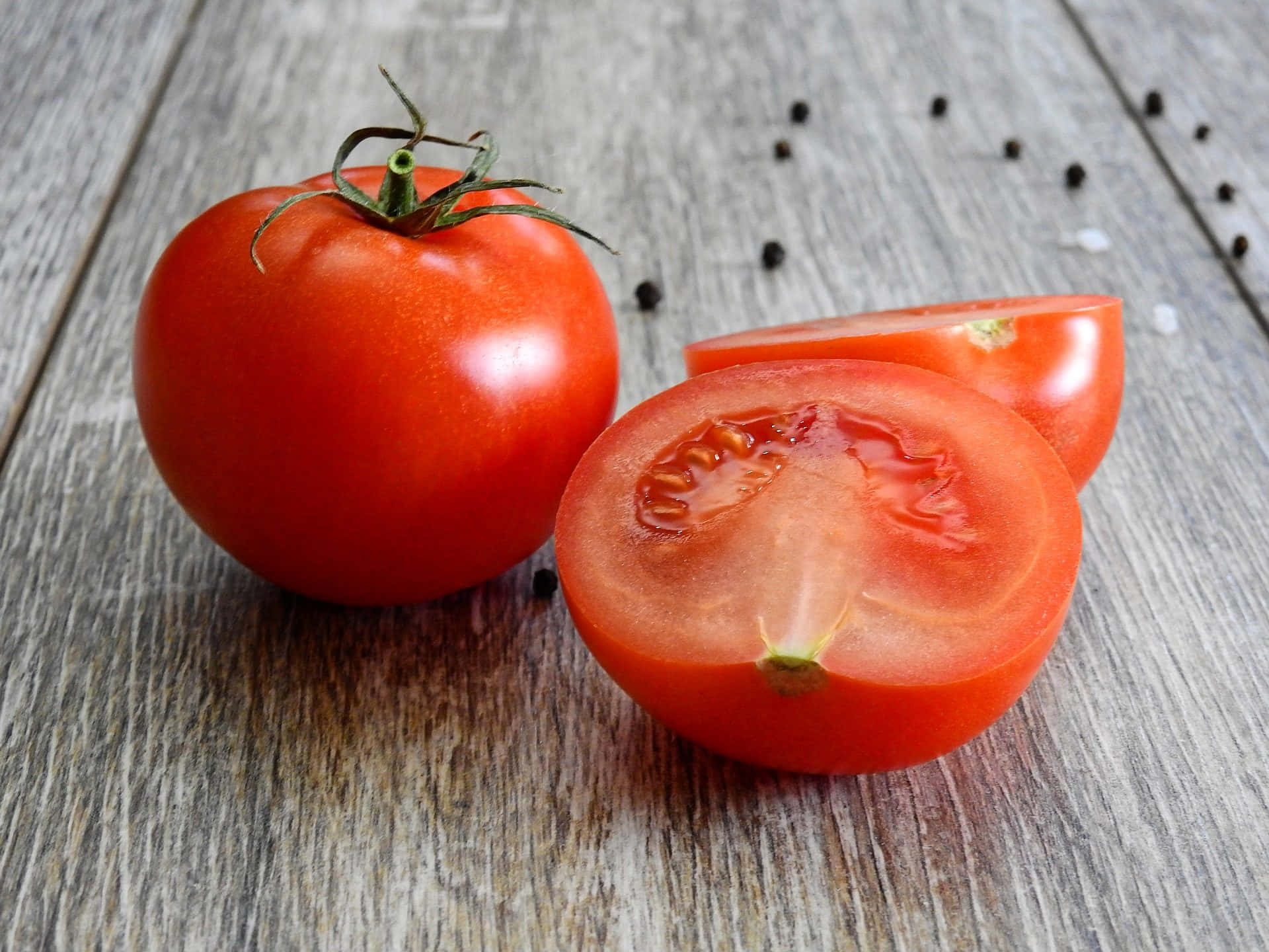 tomato 桌布 壁紙 — 一個鮮紅色的番茄，完整和切半，在風化的木質表面上露出多汁的內部，周圍散落著胡椒粒。
