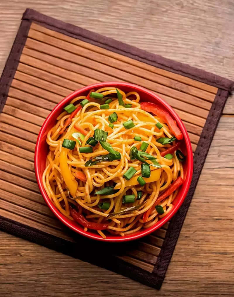 Food Photography 桌布 壁紙 — 一個鮮紅色的碗盛載著炒麵，配以色彩繽紛的甜椒和切碎的青蔥，放在木桌上的竹蓆上。