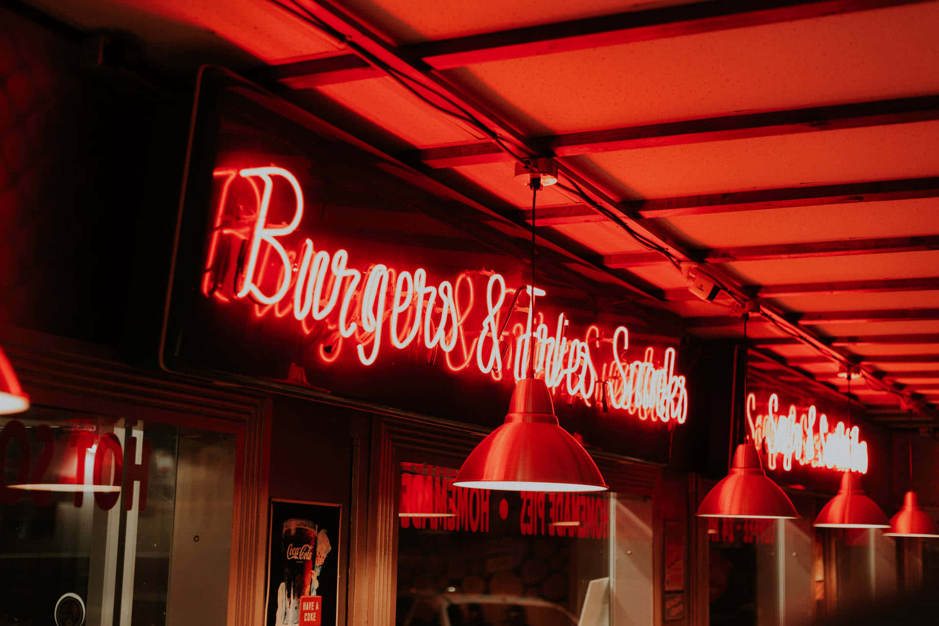 Riverdale 桌布 壁紙 — 一個鮮紅色的霓虹招牌寫著 Burgers & Fries，在夜晚的餐館入口上方溫暖地發光，將火紅的色調投射在吊燈和裸露的橫樑上。