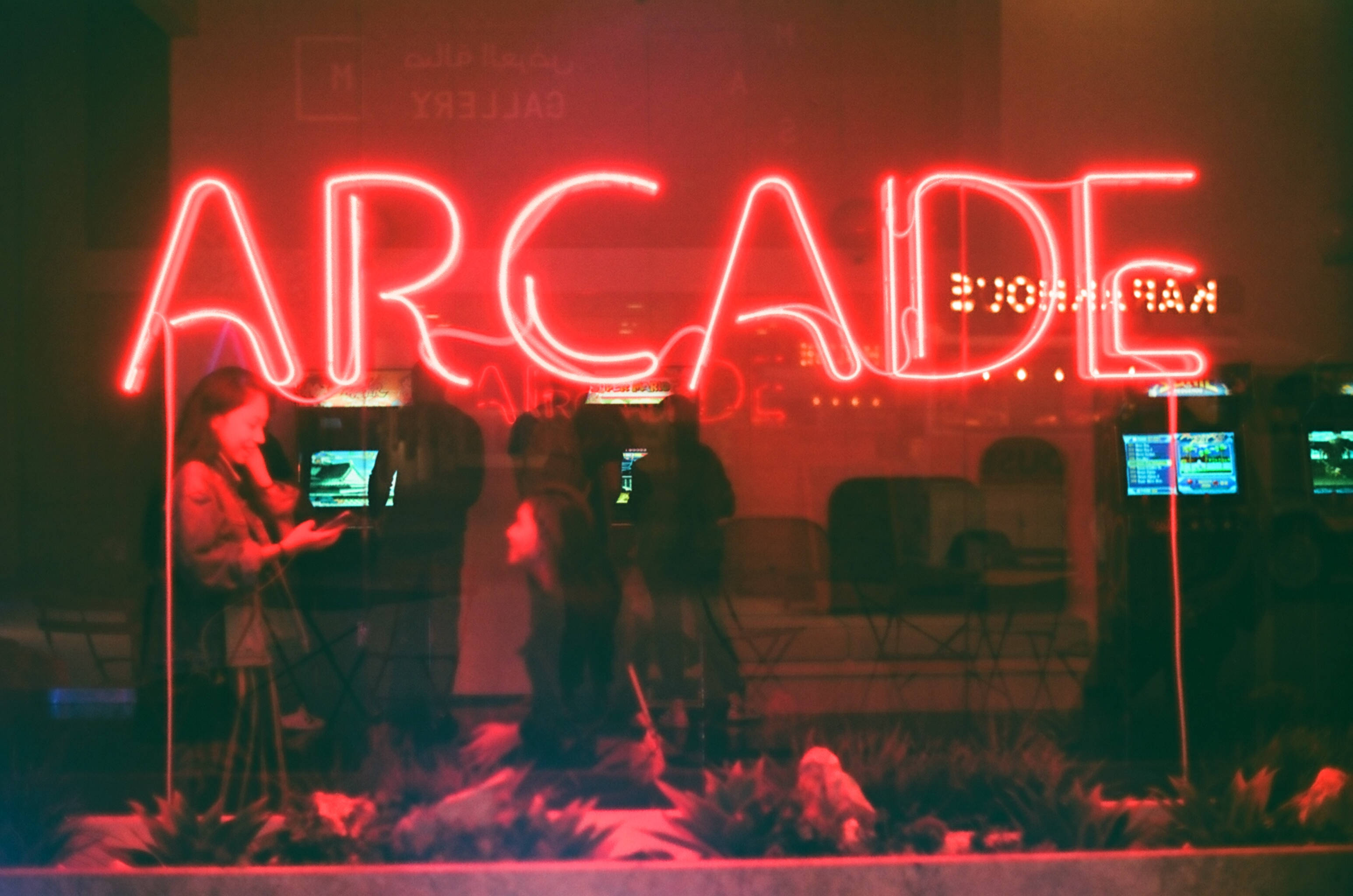 美學攝影 桌布 壁紙 — 一個鮮紅色的霓虹燈牌，拼寫著ARCADE，在昏暗的城市環境中，在玩復古遊戲的模糊人物背後發光。