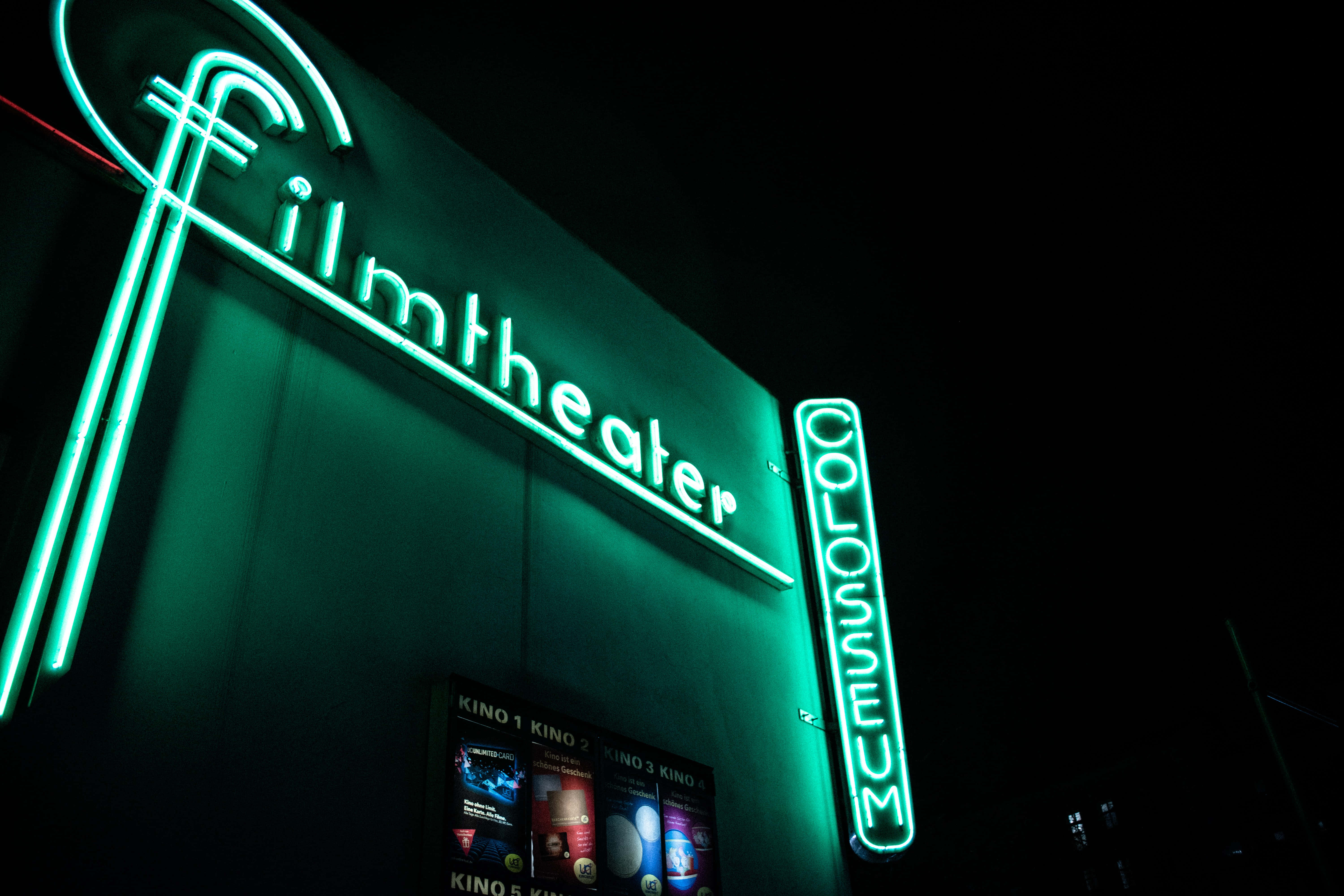 深綠色系動畫 桌布 壁紙 — 一個鮮綠色霓虹燈牌，上面寫著「Filmtheater」，在夜間照亮黑暗的建築物外牆，帶有發光字體和垂直的「CINEMA」文字。