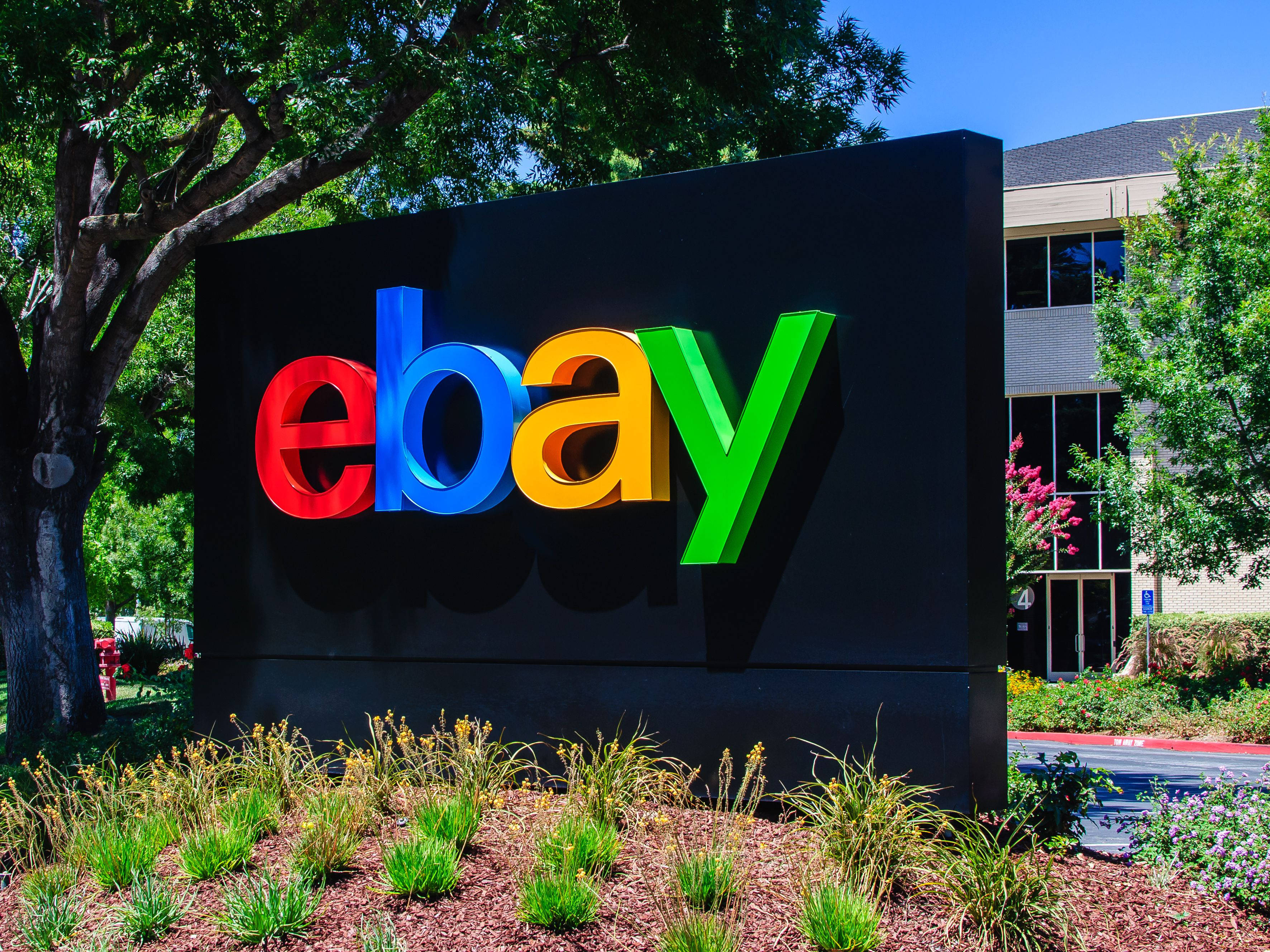 eBay 桌布 壁紙 — 一個鮮豔的eBay標誌招牌，帶有紅色、藍色、黃色和綠色的字母，矗立在戶外，在晴朗的藍天下，周圍環繞著茂密的綠樹和園林植物。