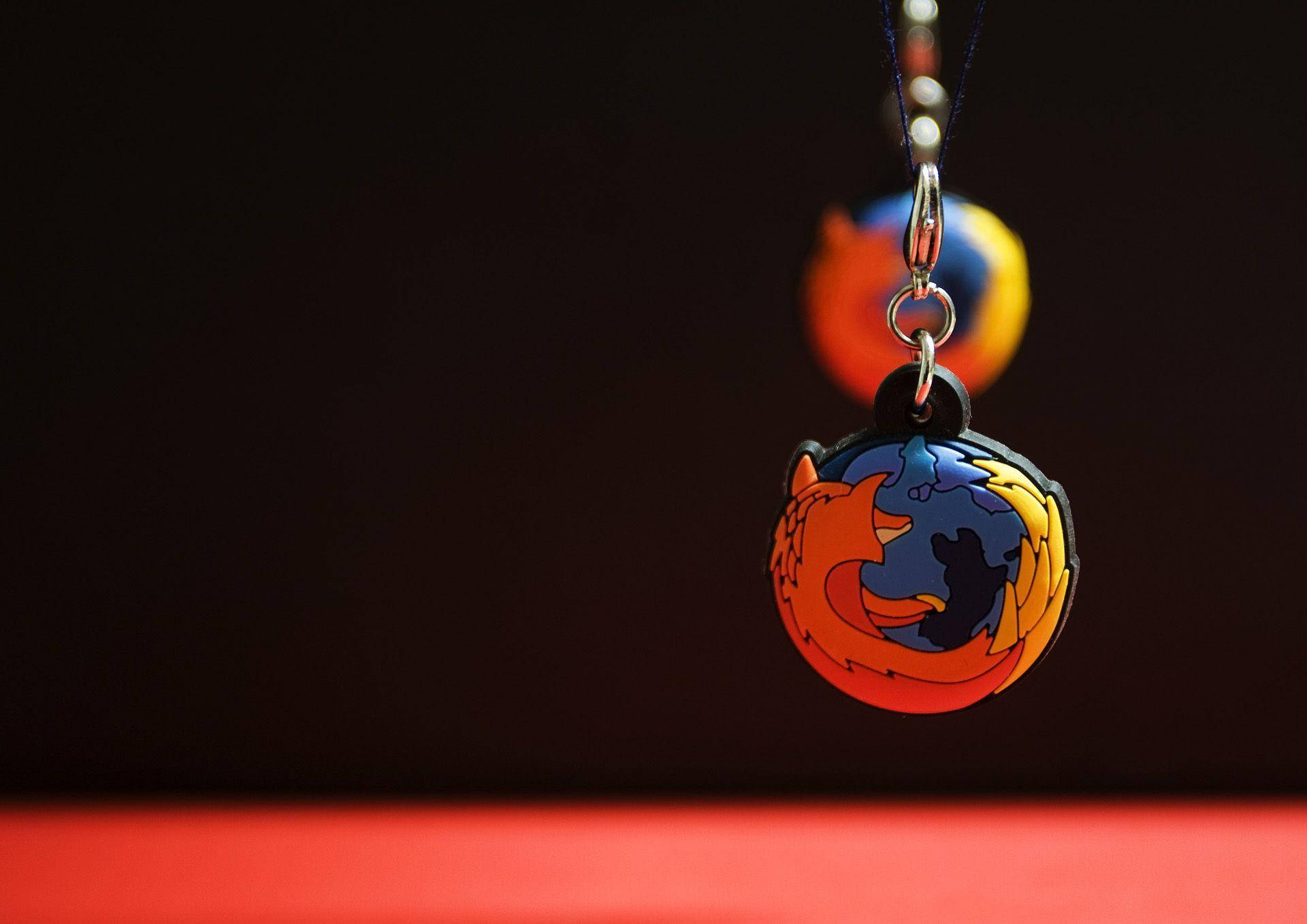 Website 桌布 壁紙 — 一個鮮豔的Firefox標誌鑰匙圈在黑暗背景中清晰懸掛，背景中有模糊的第二個鑰匙圈和下方的紅色表面。