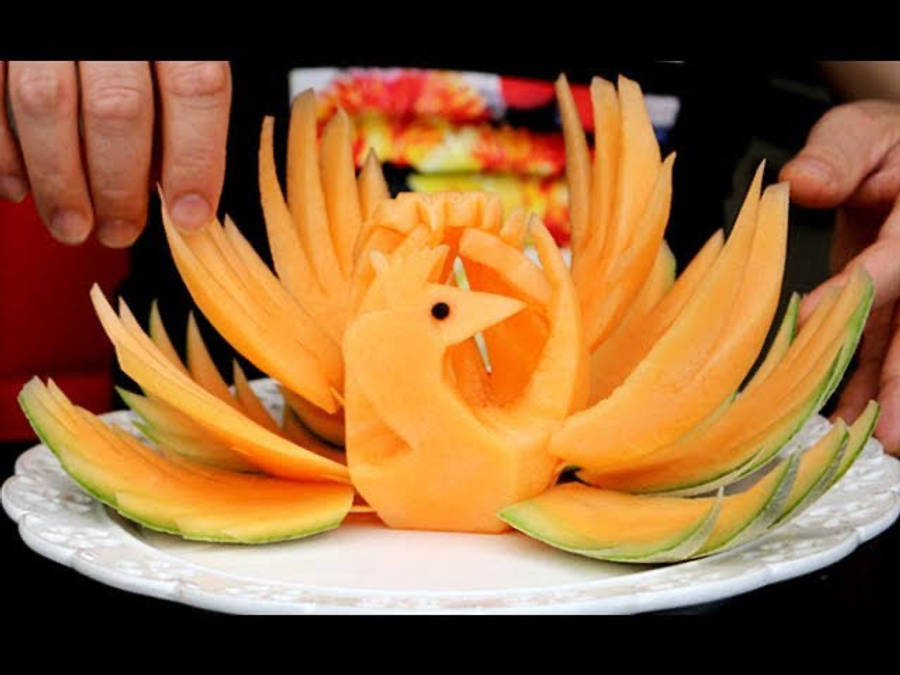 Fruit 桌布 壁紙 — 一個鮮豔的哈密瓜雕刻成精細的鳳凰，展開橙色翅膀和綠色瓜皮邊緣，放在白色盤子上。