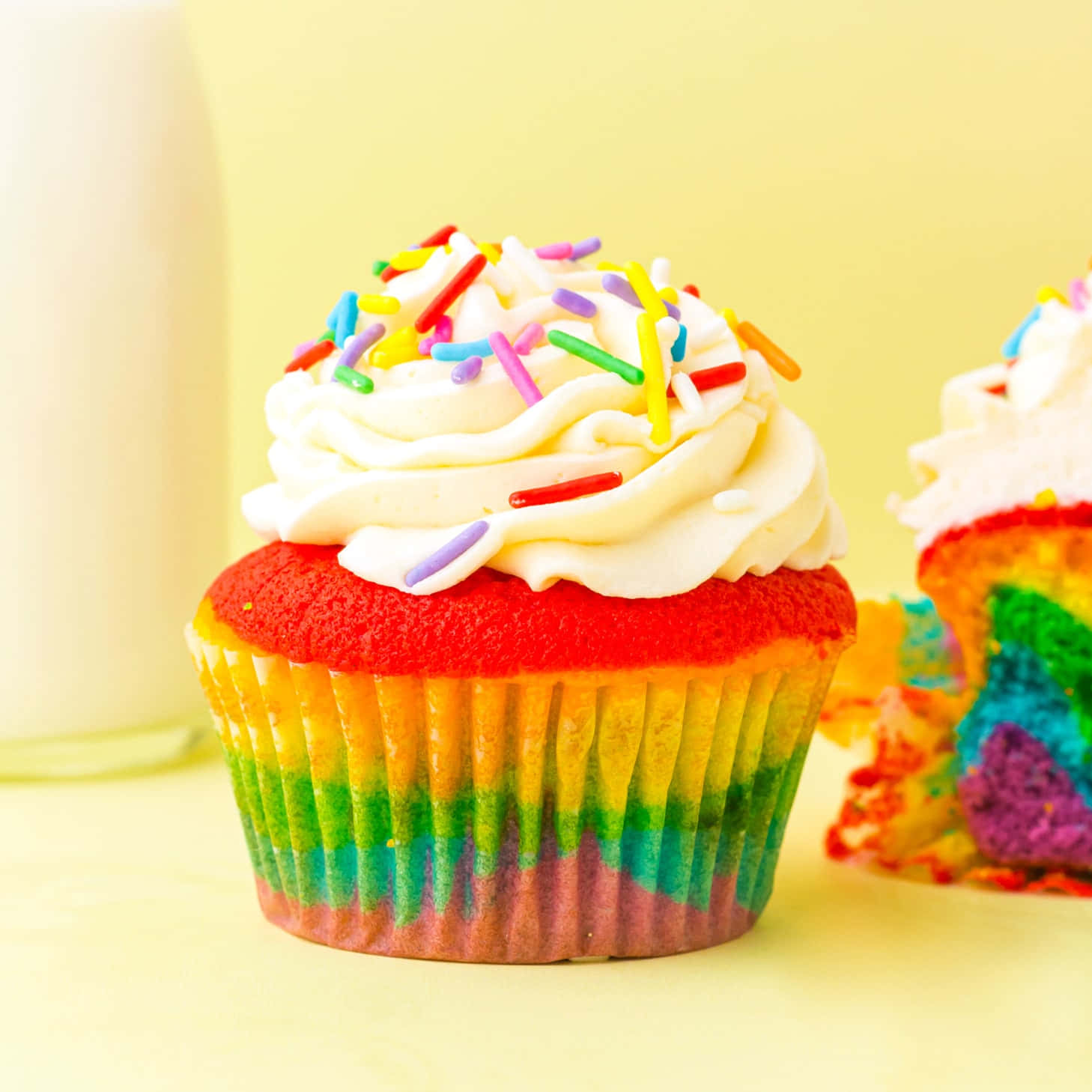 Cupcake 桌布 壁紙 — 一個鮮豔的彩虹杯子蛋糕，上面有彩色糖粒和奶油糖霜，旁邊放著一個切開的，露出層層內餡。