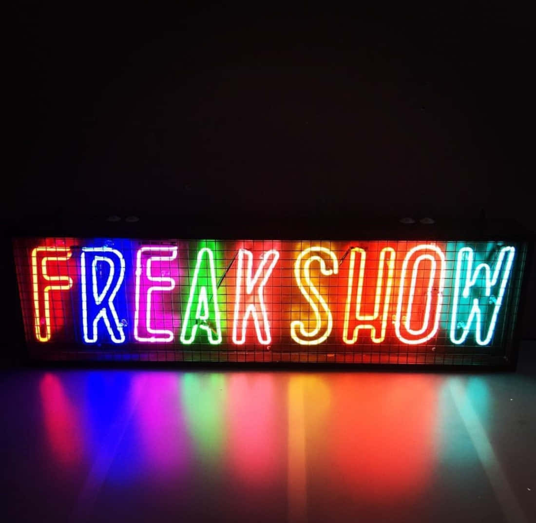霓虹美學 桌布 壁紙 — 一個鮮豔的彩虹霓虹燈牌，拼寫著 FREAK SHOW，在黑暗背景中發光，並在光滑表面上投射出色彩繽紛的倒影。