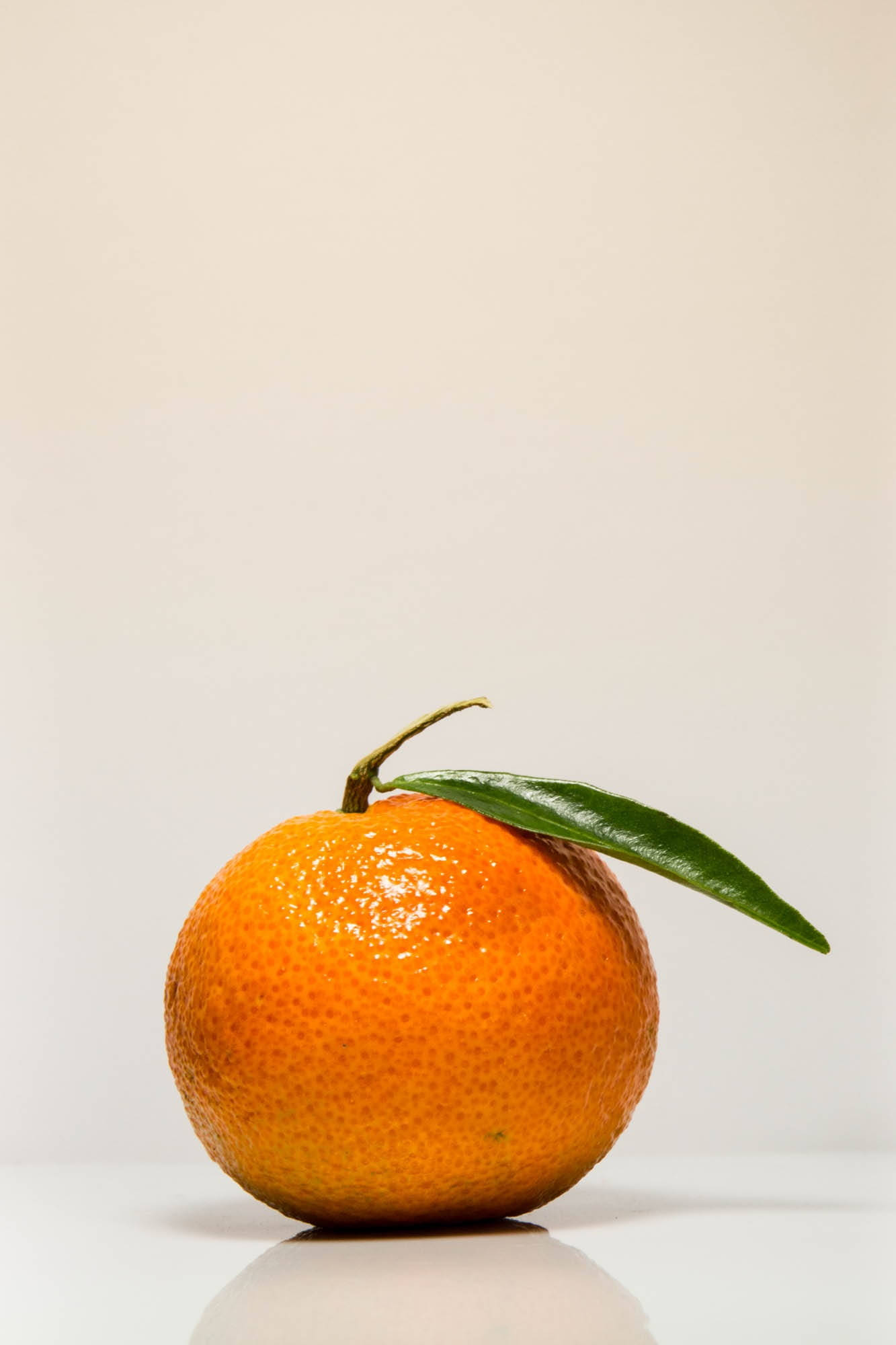 Fruit 桌布 壁紙 — 一個鮮豔的柳橙，帶有新鮮的綠葉，放在白色表面上，背景是柔和的米色。