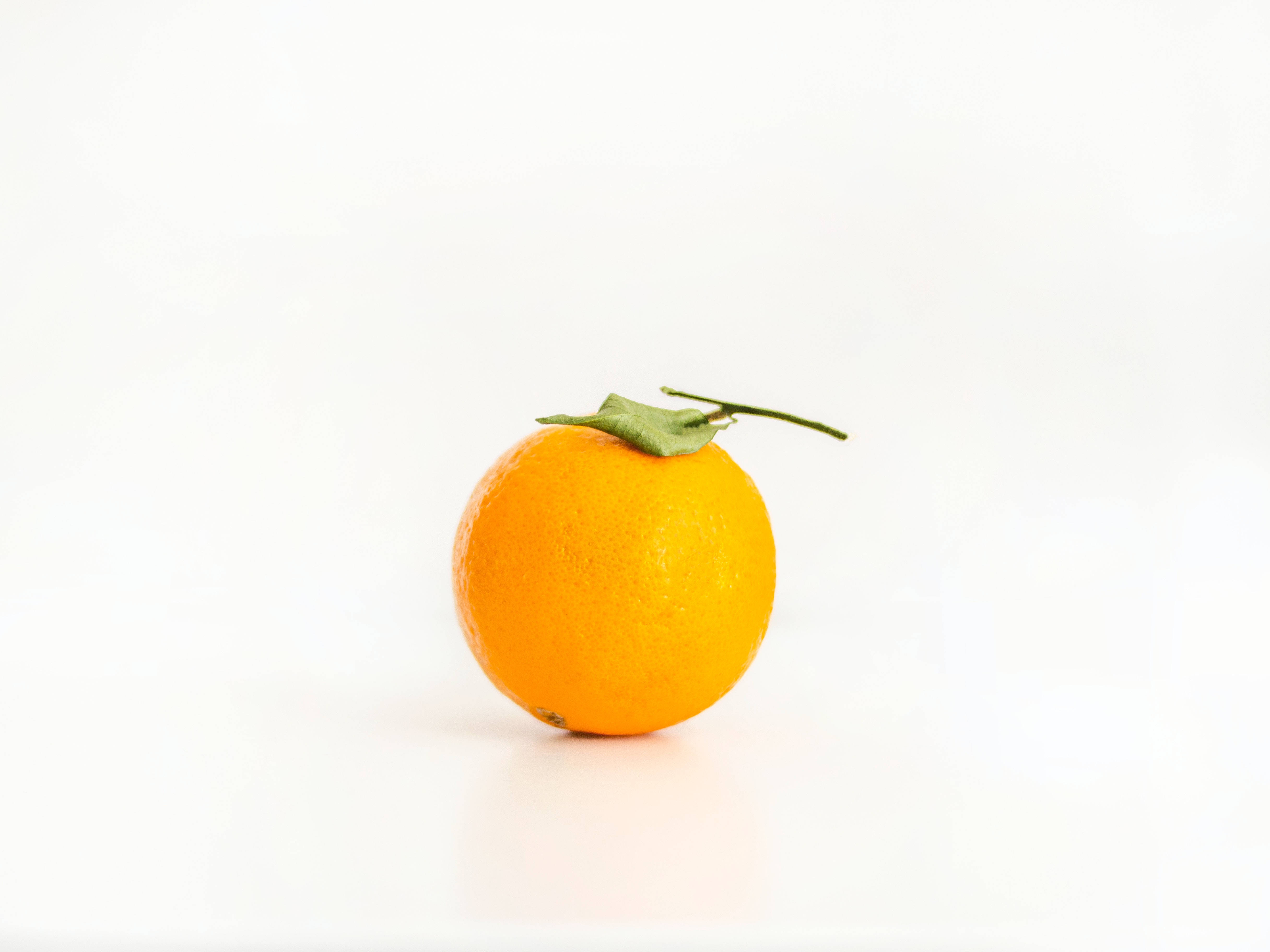 Fruit 桌布 壁紙 — 一個鮮豔的柳橙，帶有綠色莖，位於明亮的白色表面中央，投下柔和的陰影。