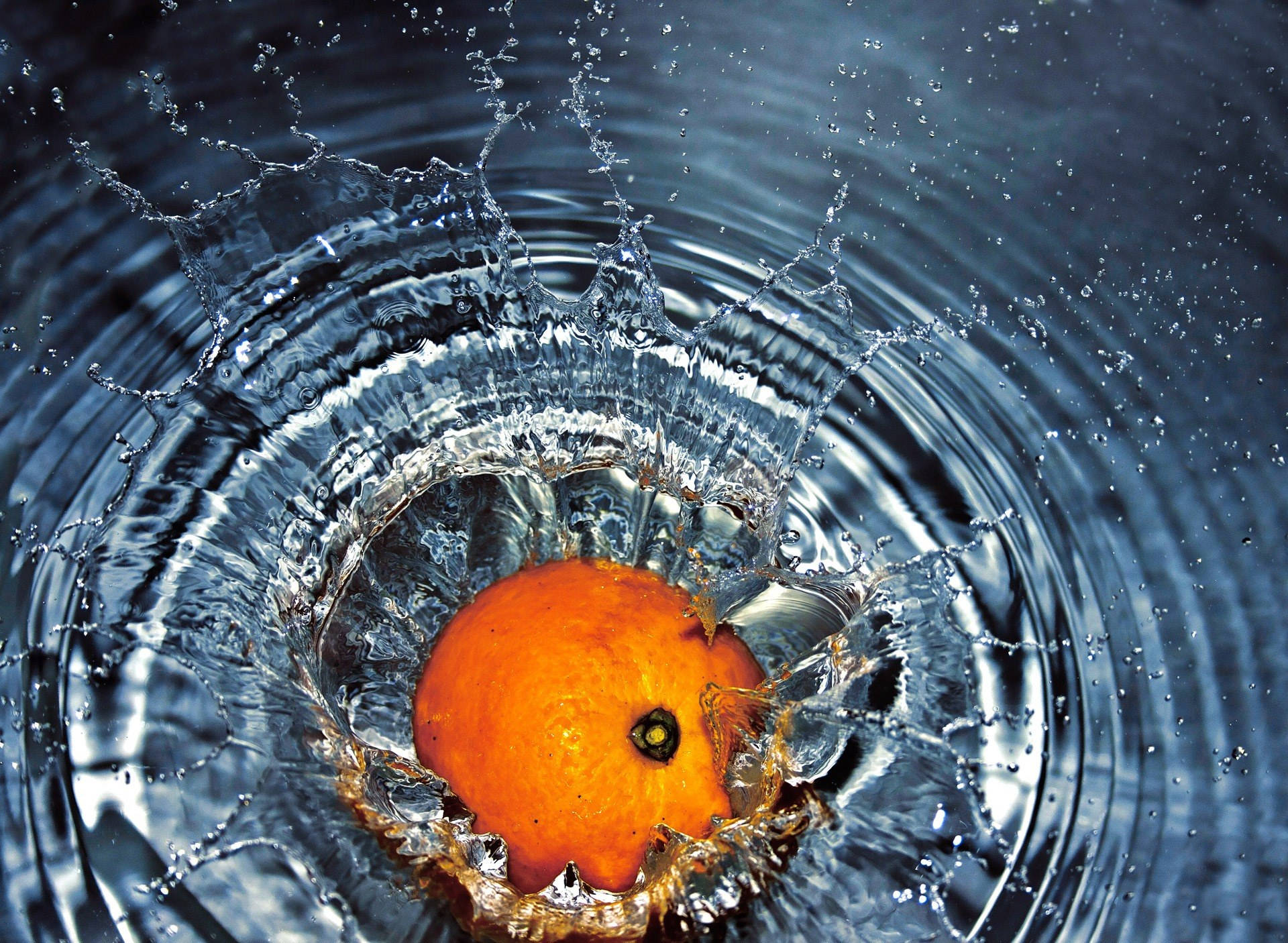 Falling Water 桌布 壁紙 — 一個鮮豔的橙子潛入深藍色的水中，產生爆炸性的漣漪和閃閃發光的水滴。