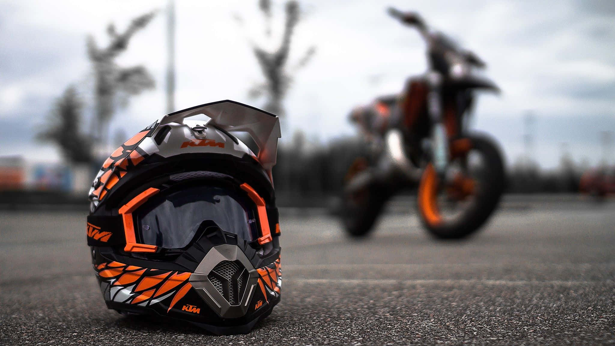 Ktm Bike 桌布 壁紙 — 一個鮮豔的橙色和黑色KTM越野摩托車頭盔放在柏油路上，背景有一輛模糊的越野摩托車，天空多雲。
