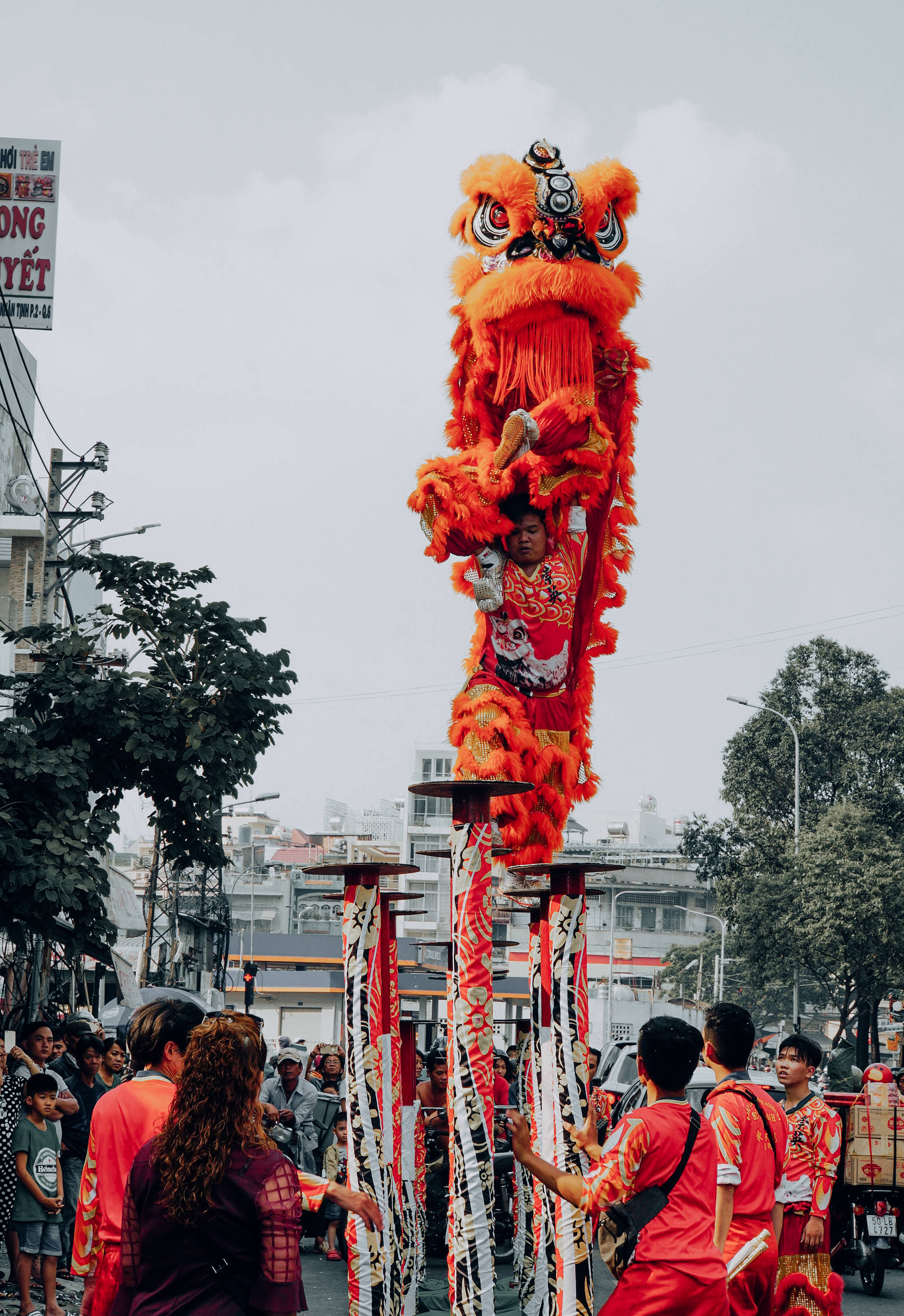 春節 桌布 壁紙 — 一個鮮豔的橙色舞獅服裝高高棲息在高桿上，街頭節日中有觀眾聚集在下方。