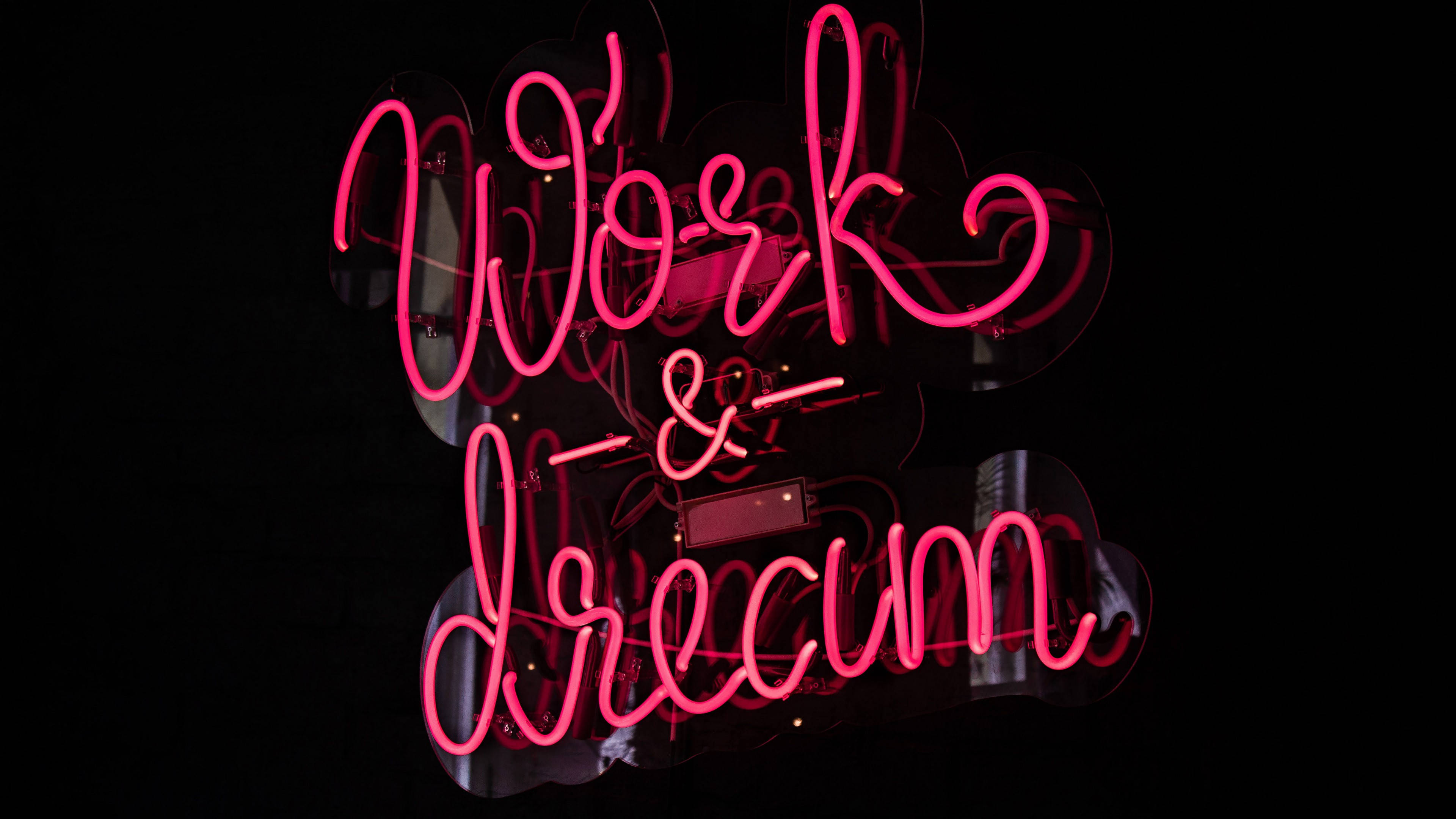 桌布 壁紙 — 一個鮮豔的粉紅色霓虹燈牌，上面寫著「Work & Dream」，在黑暗的反光表面上發光。