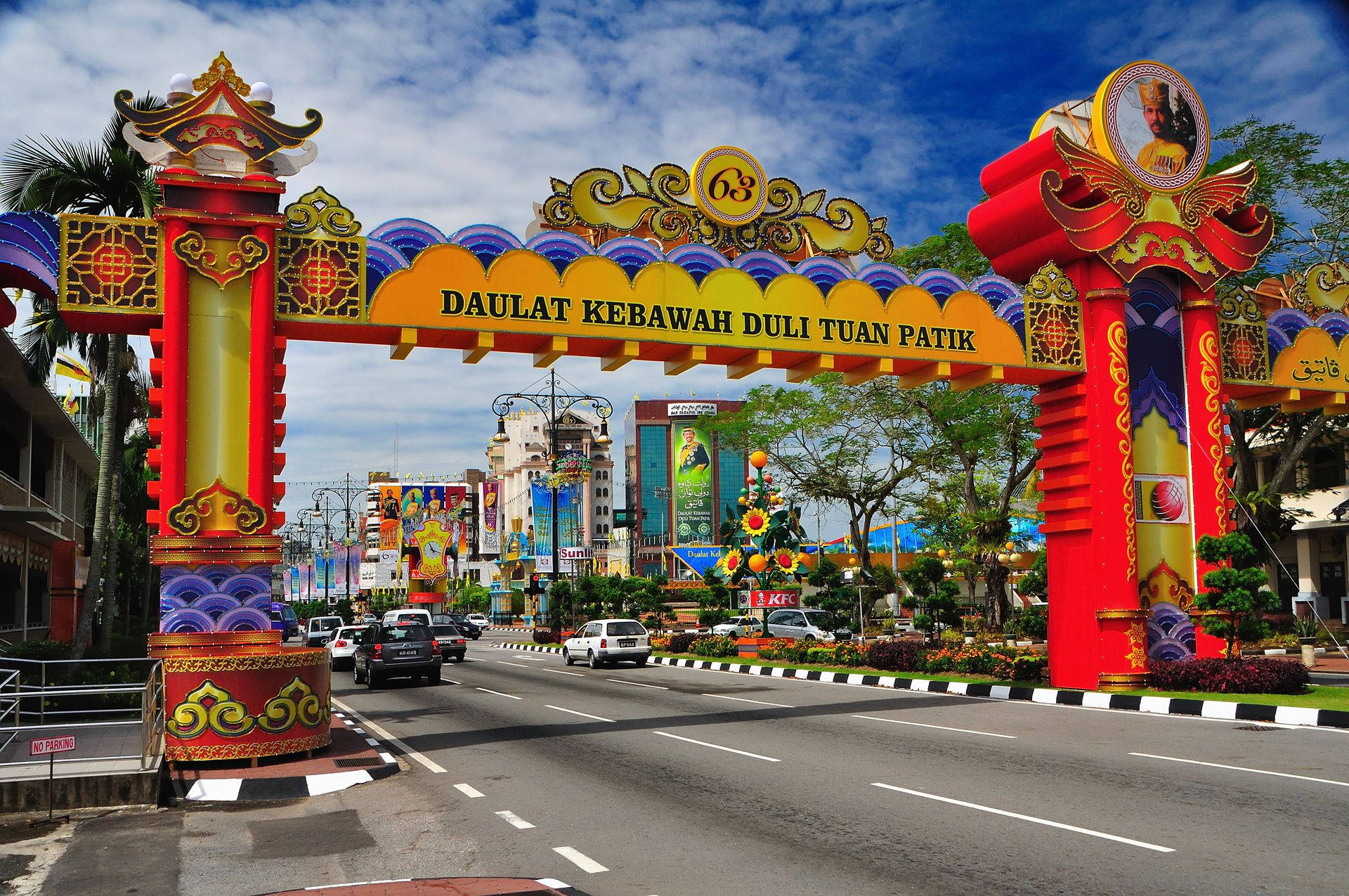 Road 桌布 壁紙 — 一個鮮豔的紅金色華麗拱門，上面有「DAULAT KEBAWAH DULI TUAN PATIK」標牌，矗立在一條繁忙的街道上，兩旁種滿棕櫚樹和色彩繽紛的建築物，在明亮的藍天下。