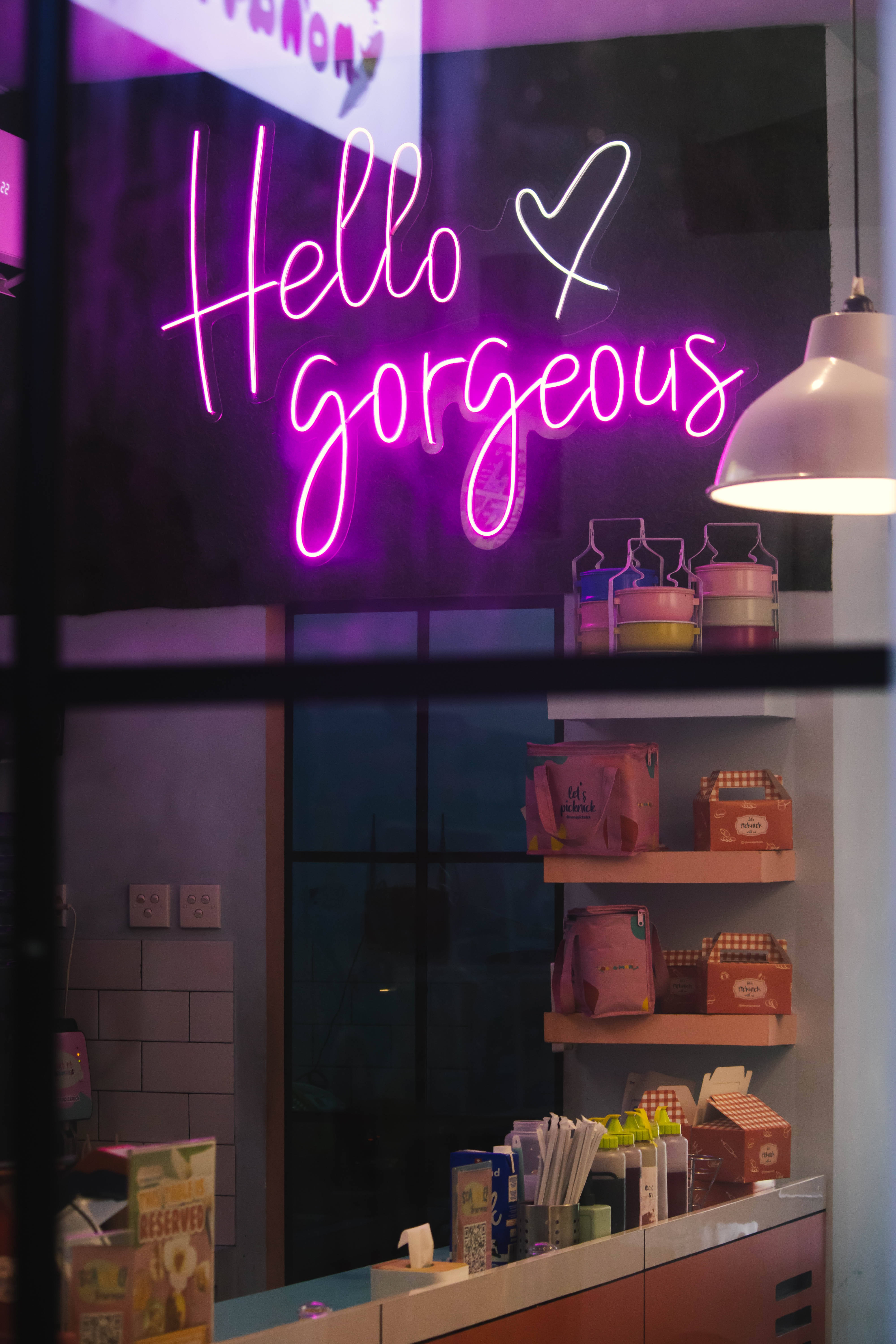 霓虹紫 iPhone 桌布 壁紙 — 一個鮮豔的紫色霓虹燈牌寫著「Hello gorgeous」，在夜間透過窗戶看到舒適的咖啡館內溫暖地發光。