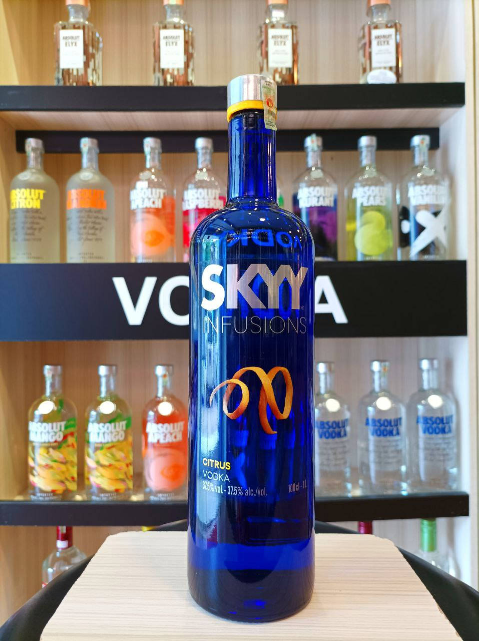 Vodka 桌布 壁紙 — 一個鮮豔的藍色 Sky Infusions Citrus 伏特加酒瓶醒目地放在木製櫃檯上，後面是擺滿色彩繽紛的 Absolut 伏特加酒瓶的架子。